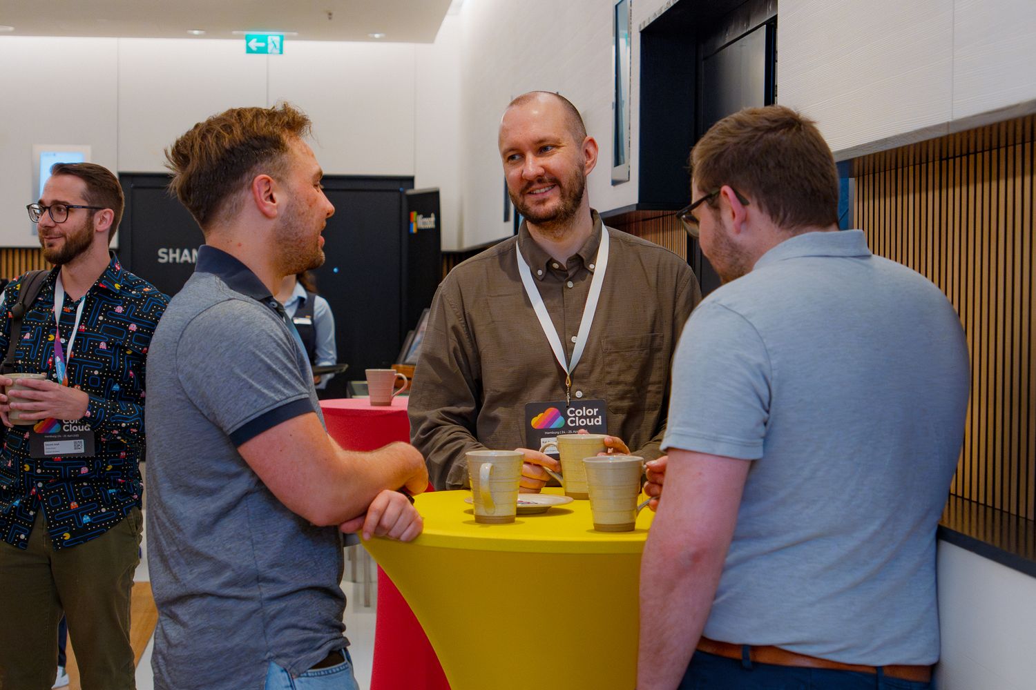 Drei Teilnehmer an der ColorCloud Konferenz stehen an einem gelben Stehtisch im Foyer und unterhalten sich