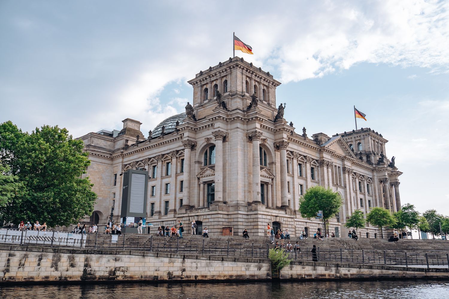 Reichstagsgebäude in Berlin mit Flaggen auf den Türmen, breiter Treppenanlage zum Fluss und vielen Menschen am Ufer