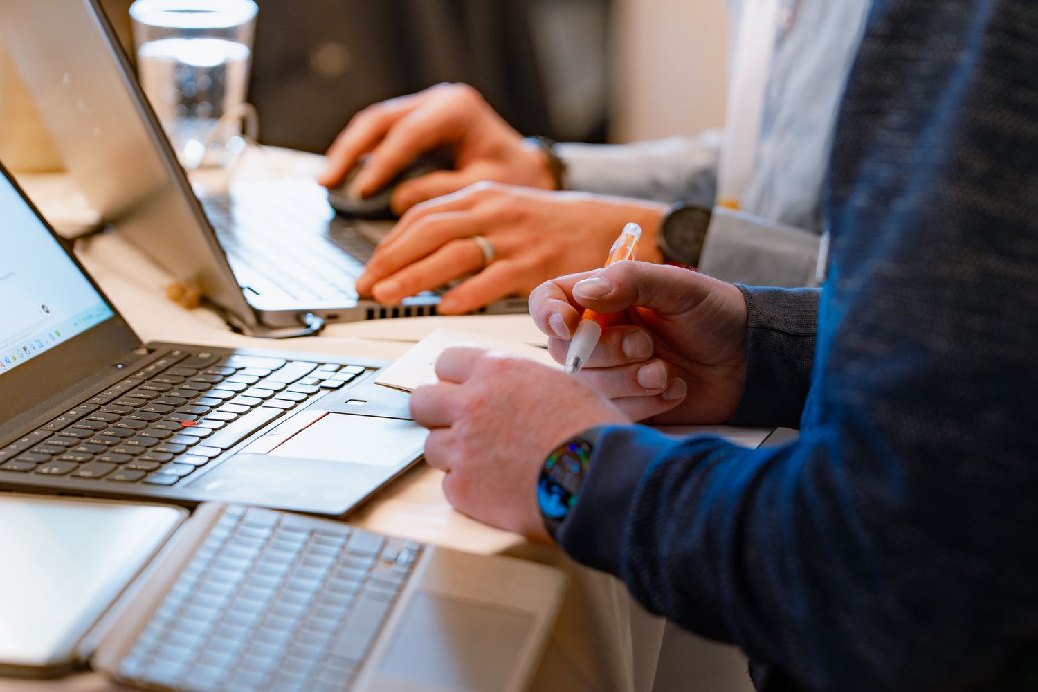 Hände mehrerer Workshopteilnehmender an Laptops, eine Person hält einen Stift über einem Notizblock