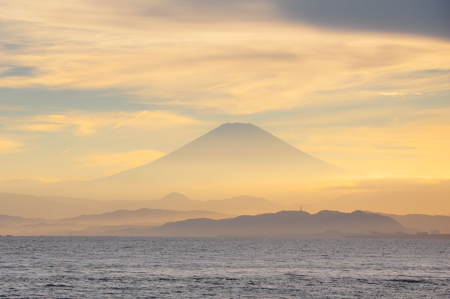Der japanische Berg Fuji-san erhebt sich im Dunst über dem Meer bei Sonnenuntergang, mit sanften orange-blauen Farbübergängen am Himmel