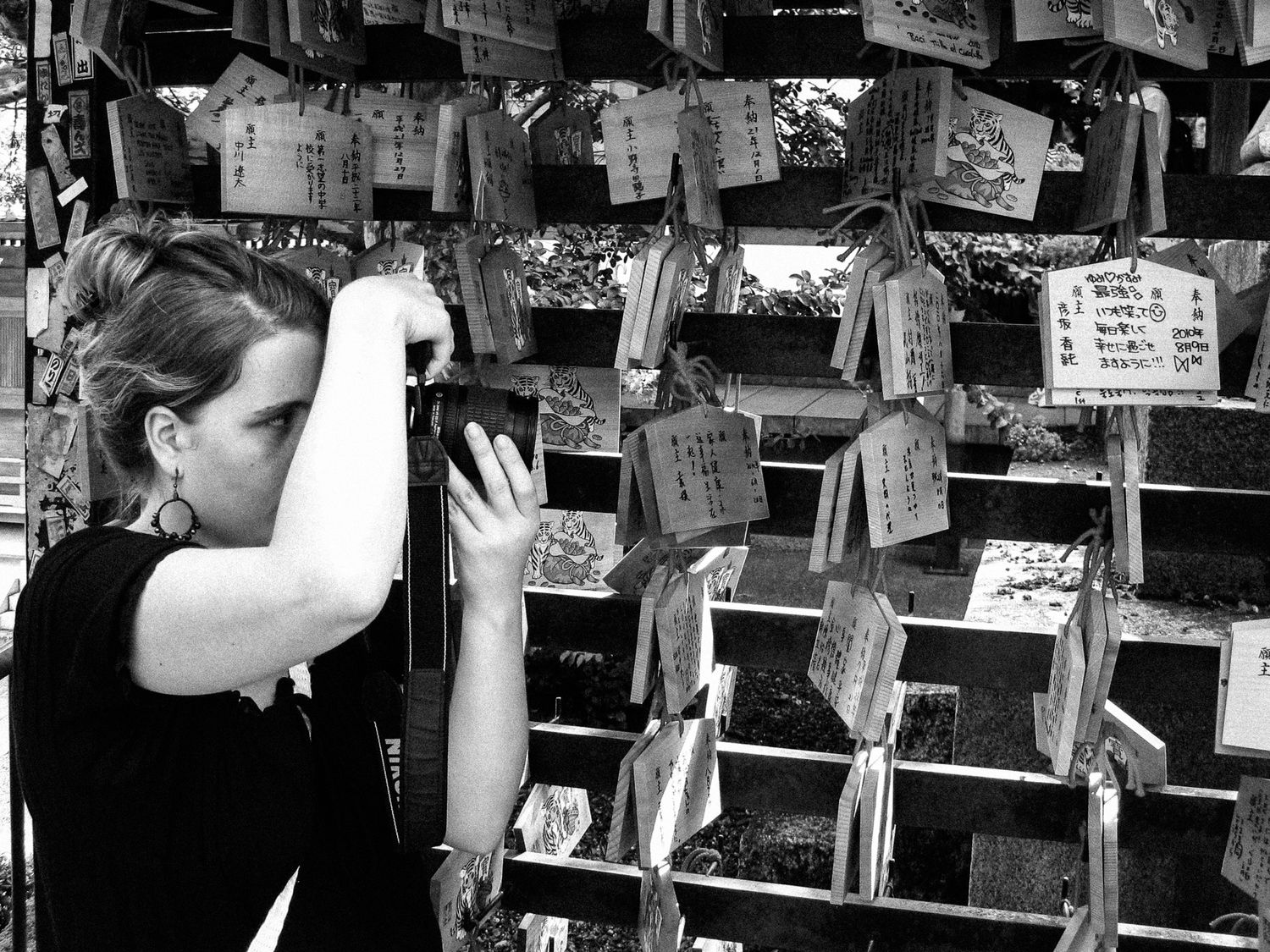 Eine junge Frau steht vor aufgehängten ema, japanische Holztafeln, und schaut durch den Sucher ihrer Nikon-Kamera