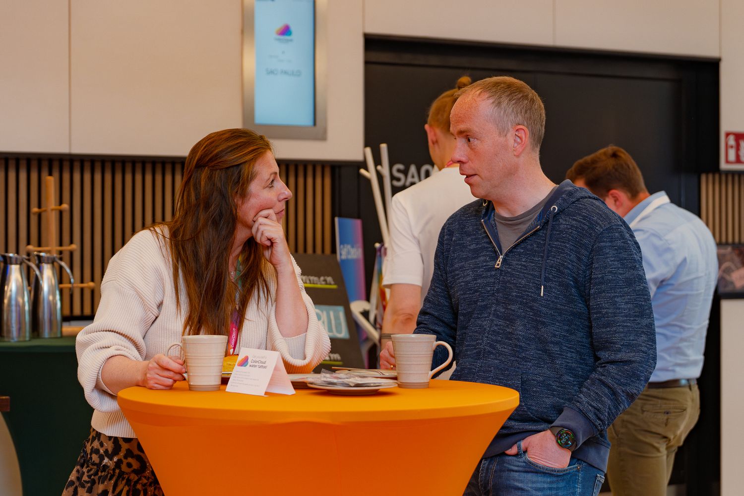 Eine Frau und ein Mann unterhalten sich angeregt an einem orangenen Stehtisch im Foyer der ColorCloud-Konferenz 2025