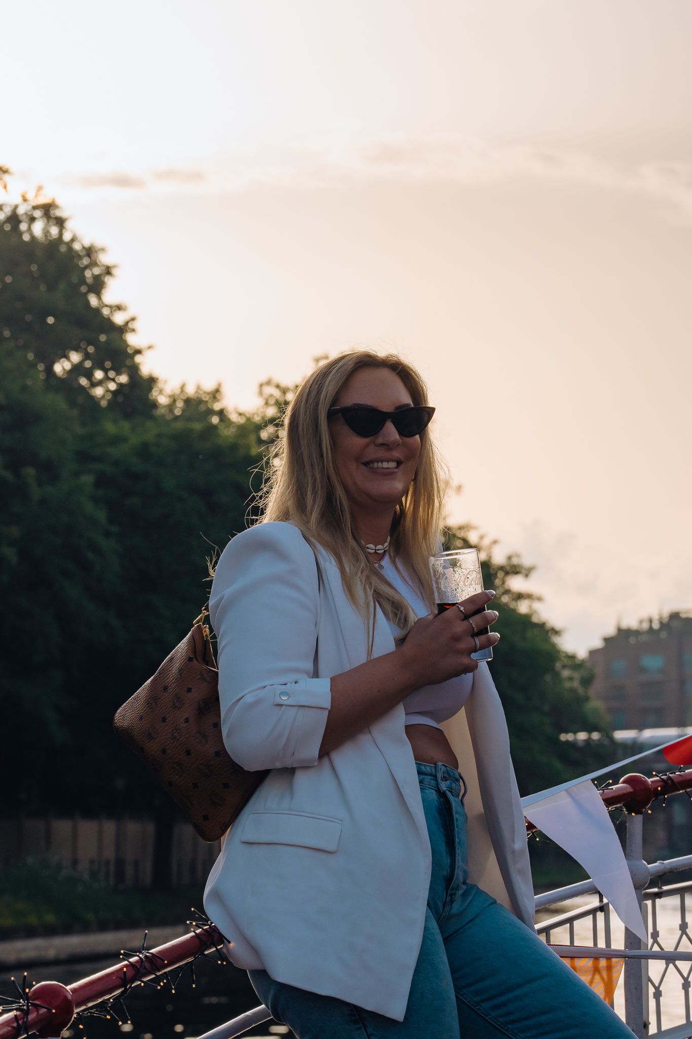 Junge Frau mit Sonnenbrille im Gegenlicht der Sonne steht an Schiffsreling gelehnt und hält ein Weinglas