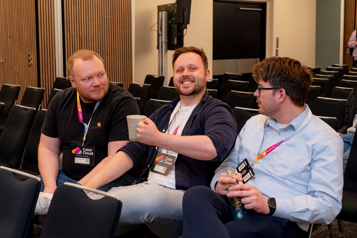 Drei Teilnehmer an der ColorCloud-Konferenz 2025 sitzen im Vortragssaal, einer hält seine Kaffeetasse hoch und grinst in die Kamera