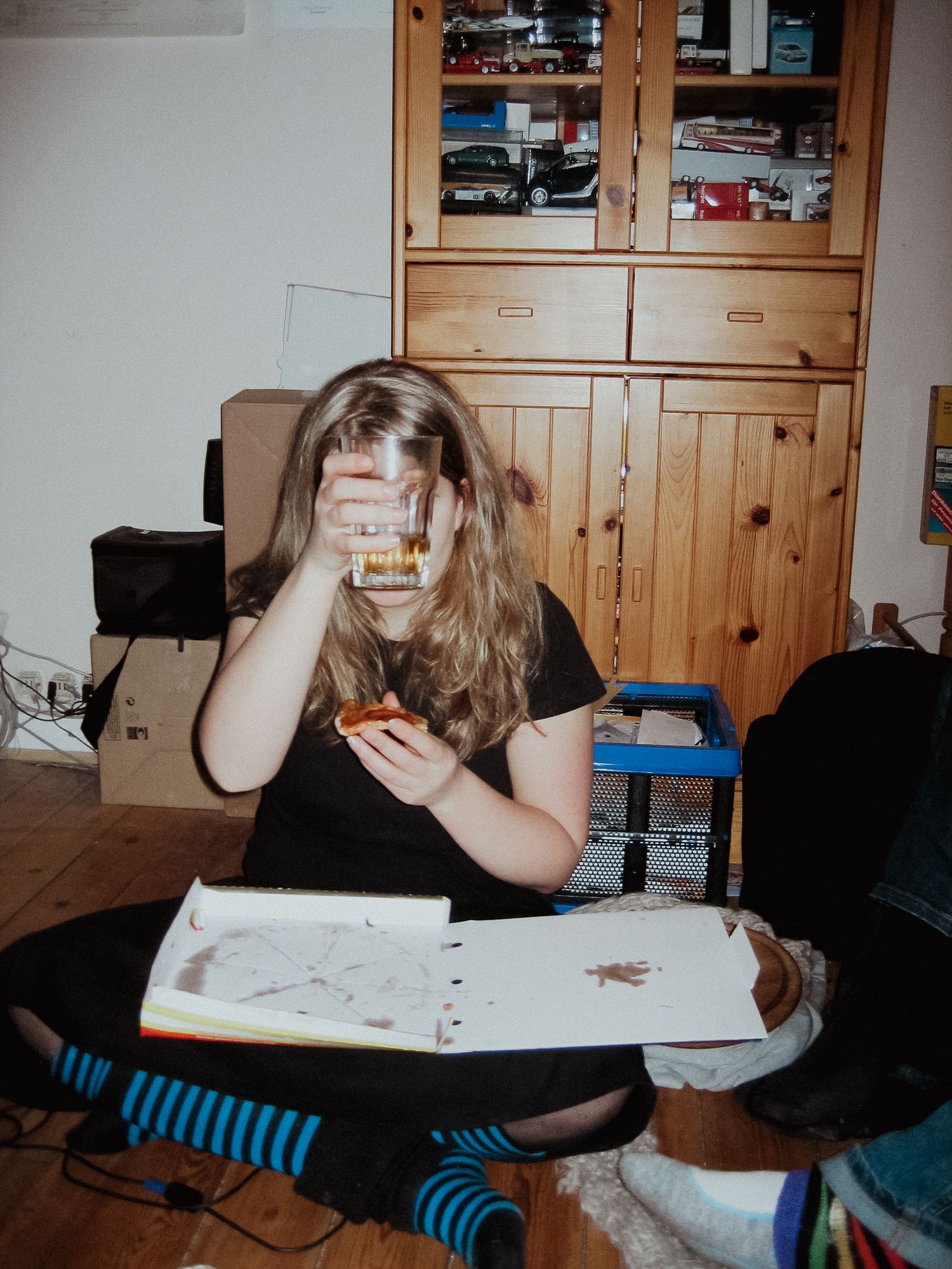 Ein Teenager mit langen blonden Haaren hält sich ein Trinkglas vor das Gesicht, in der anderen hält sie ein angebissenes Stück Pizza