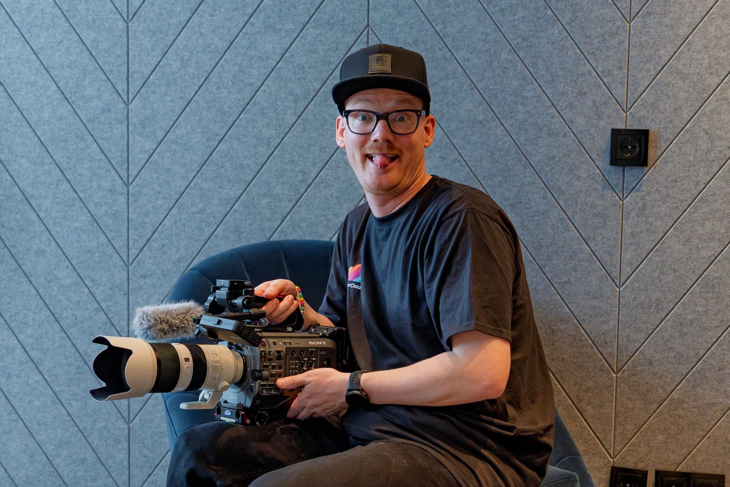 Kameramann Mario Wöhrmann hält sitzend eine große Filmkamera in der Hand und macht ein lustiges Gesicht in die Kamera