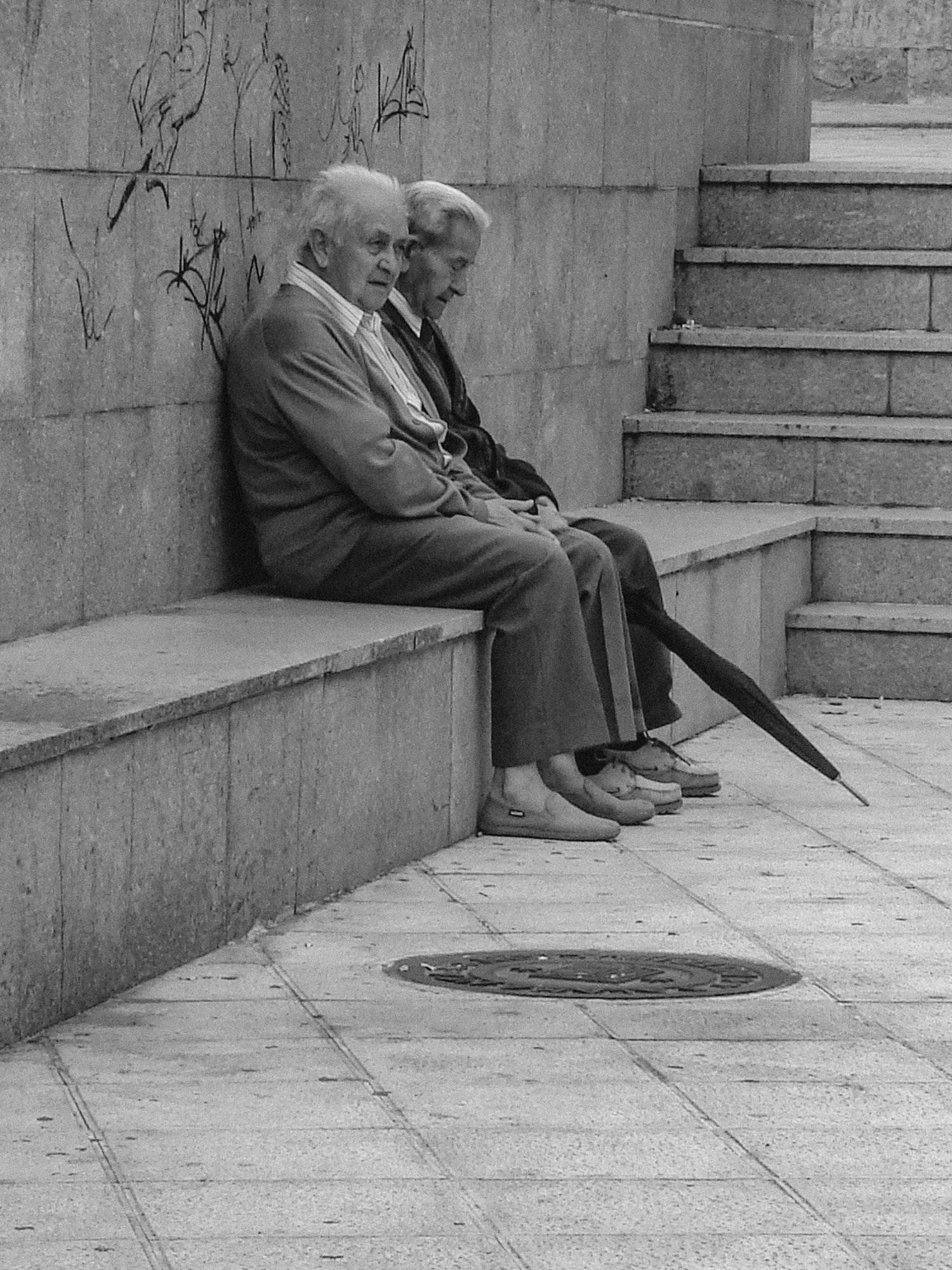 Zwei ältere Männer sitzen auf einer steinernen Treppe in Salamanca; einer hält einen schwarzen Schirm in der Hand