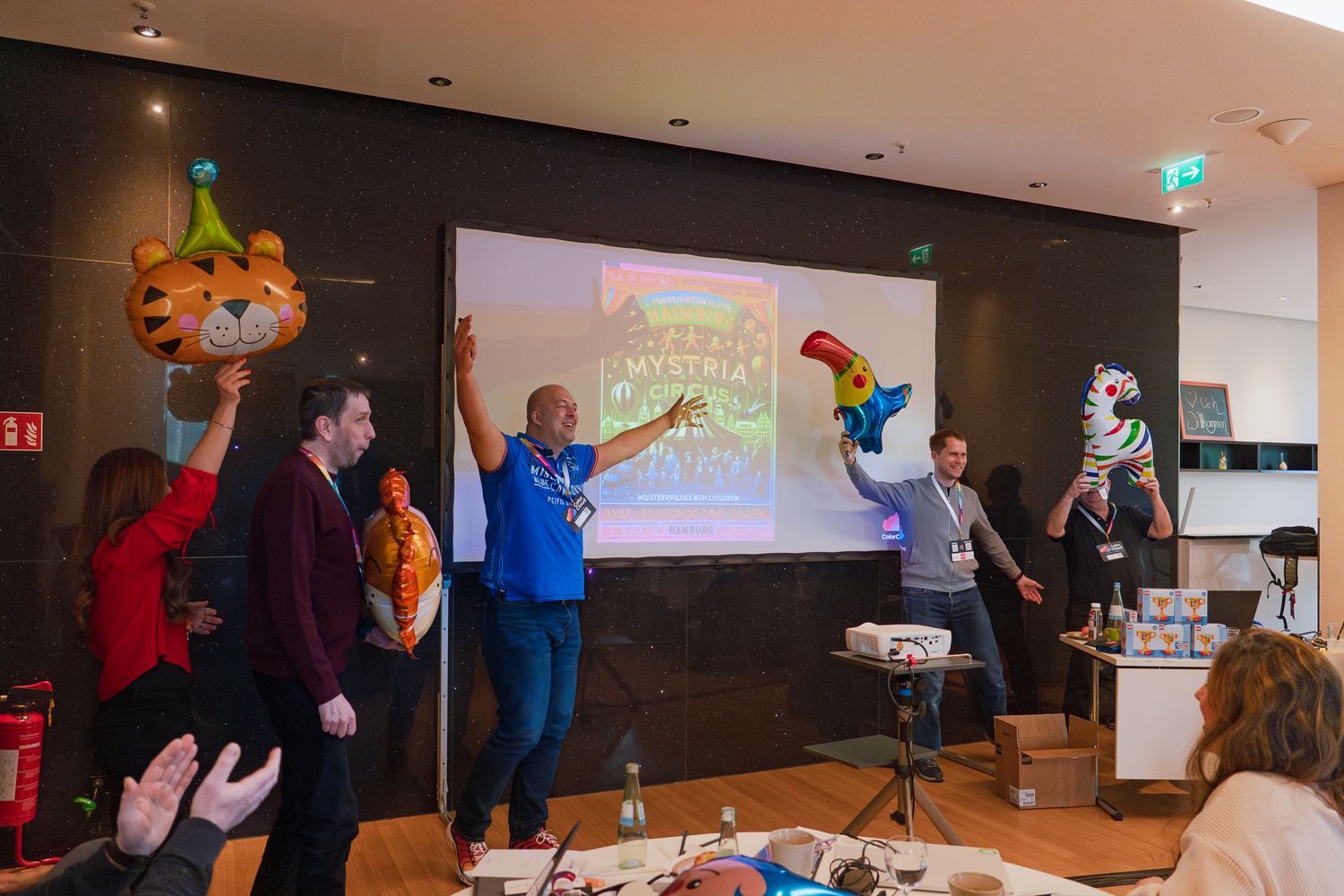 Ein Team präsentiert beim ColorCloud PromptAthon Ergebnisse vor anderen Teilnehmenden und hält dabei bunte Luftballons hoch