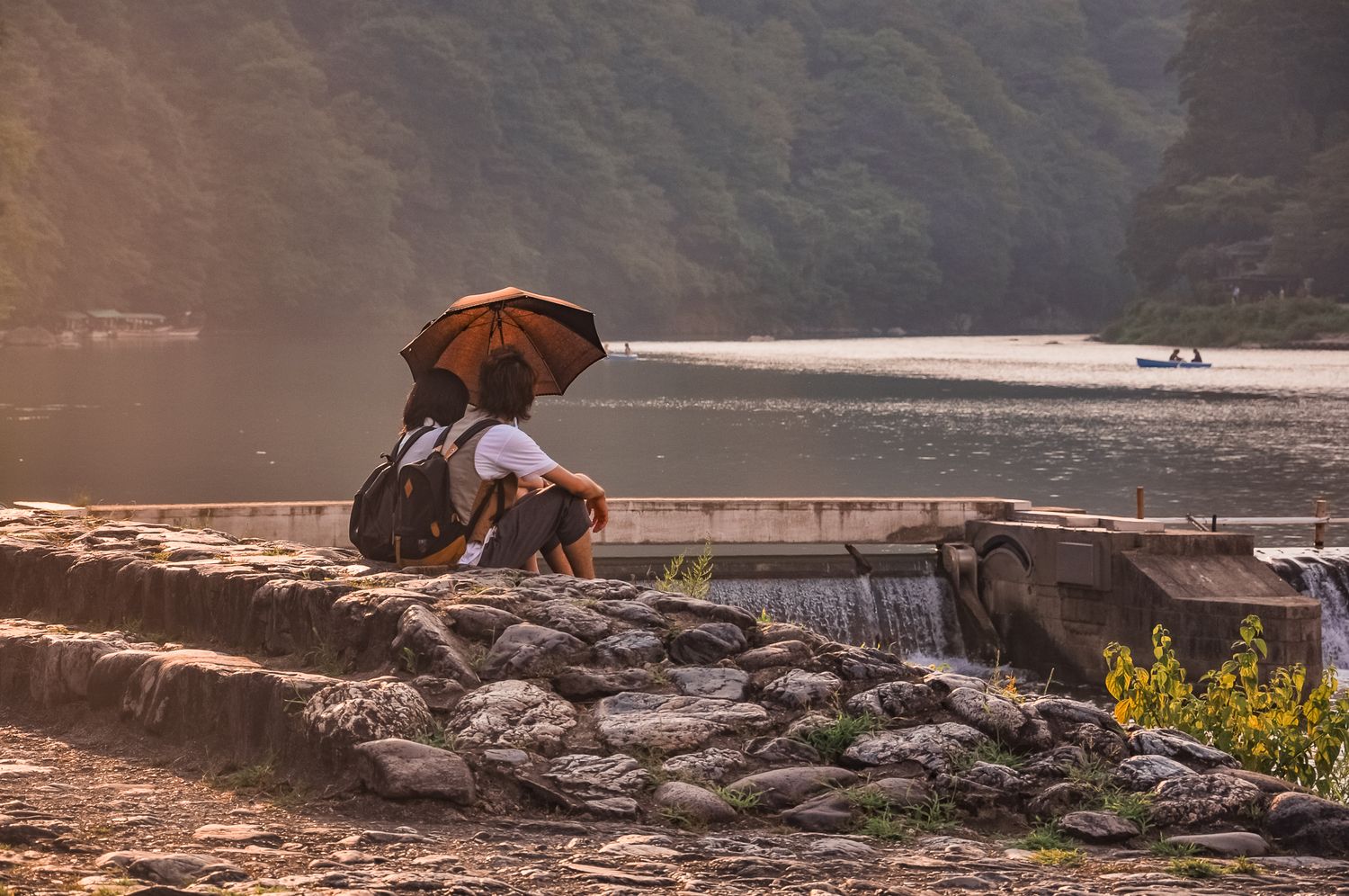 Ein Pärchen sitzt gemeinsam unter einem Sonnenschirm und blickt von einem steinernen Ufer auf den Fluss Katsura in Arashiyama