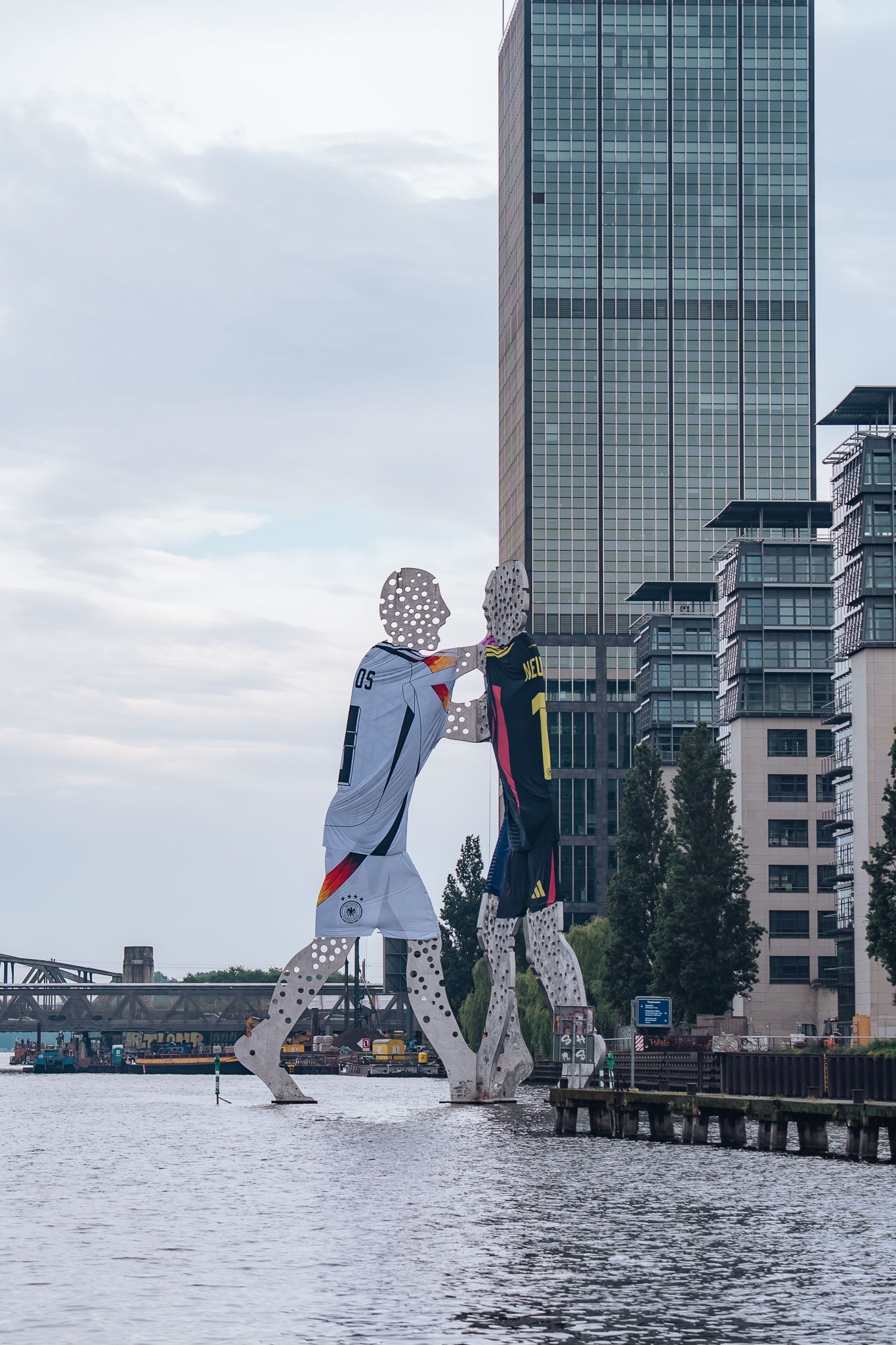 Skulptur ‚Molecule Man‘ von Jonathan Borofsky in der Spree, zwei perforierte Figuren in Fußballtrikots vor modernen Gebäuden