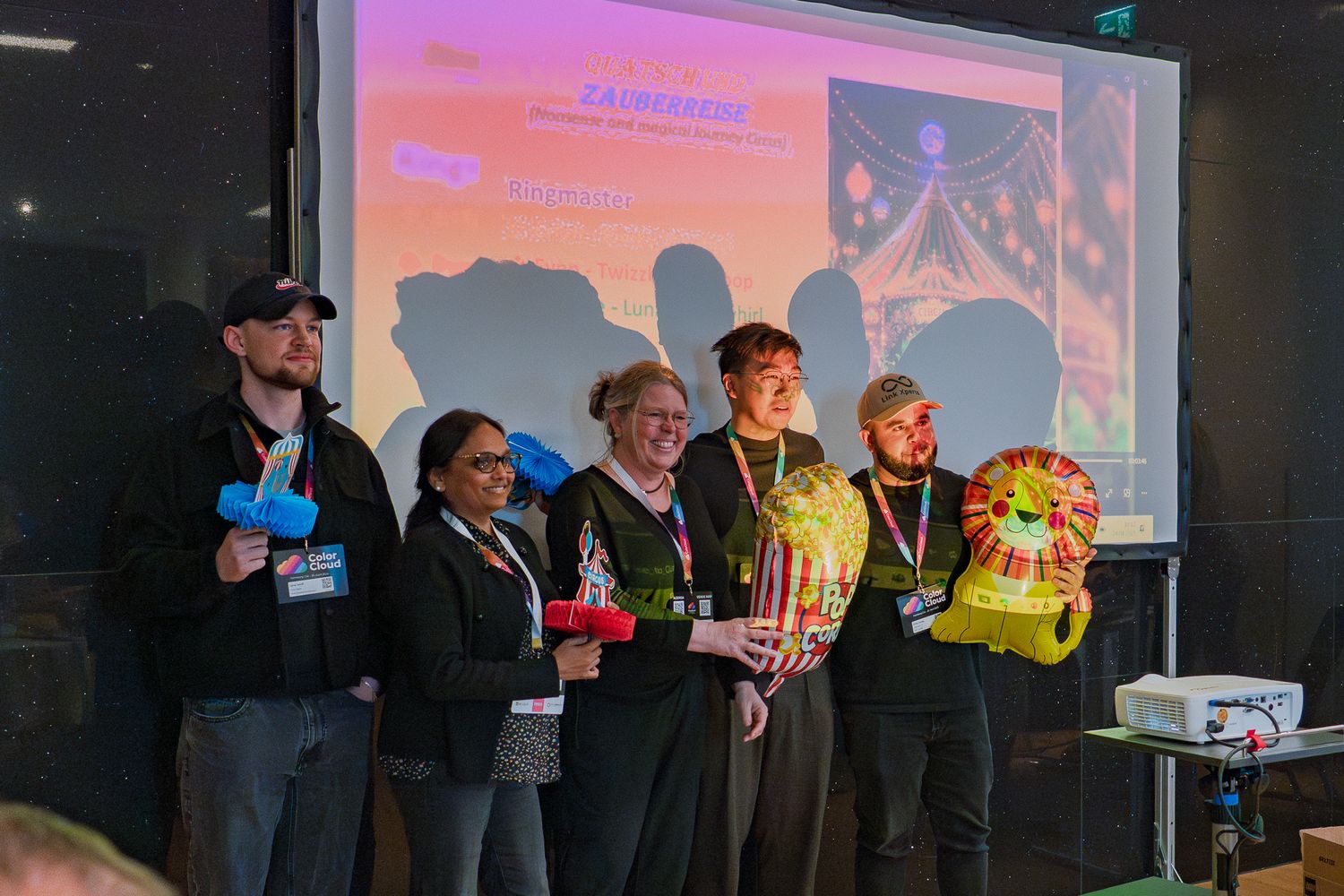 Ein Team präsentiert beim ColorCloud PromptAthon Ergebnisse vor anderen Teilnehmenden und hält dabei bunte Luftballons in der Hand