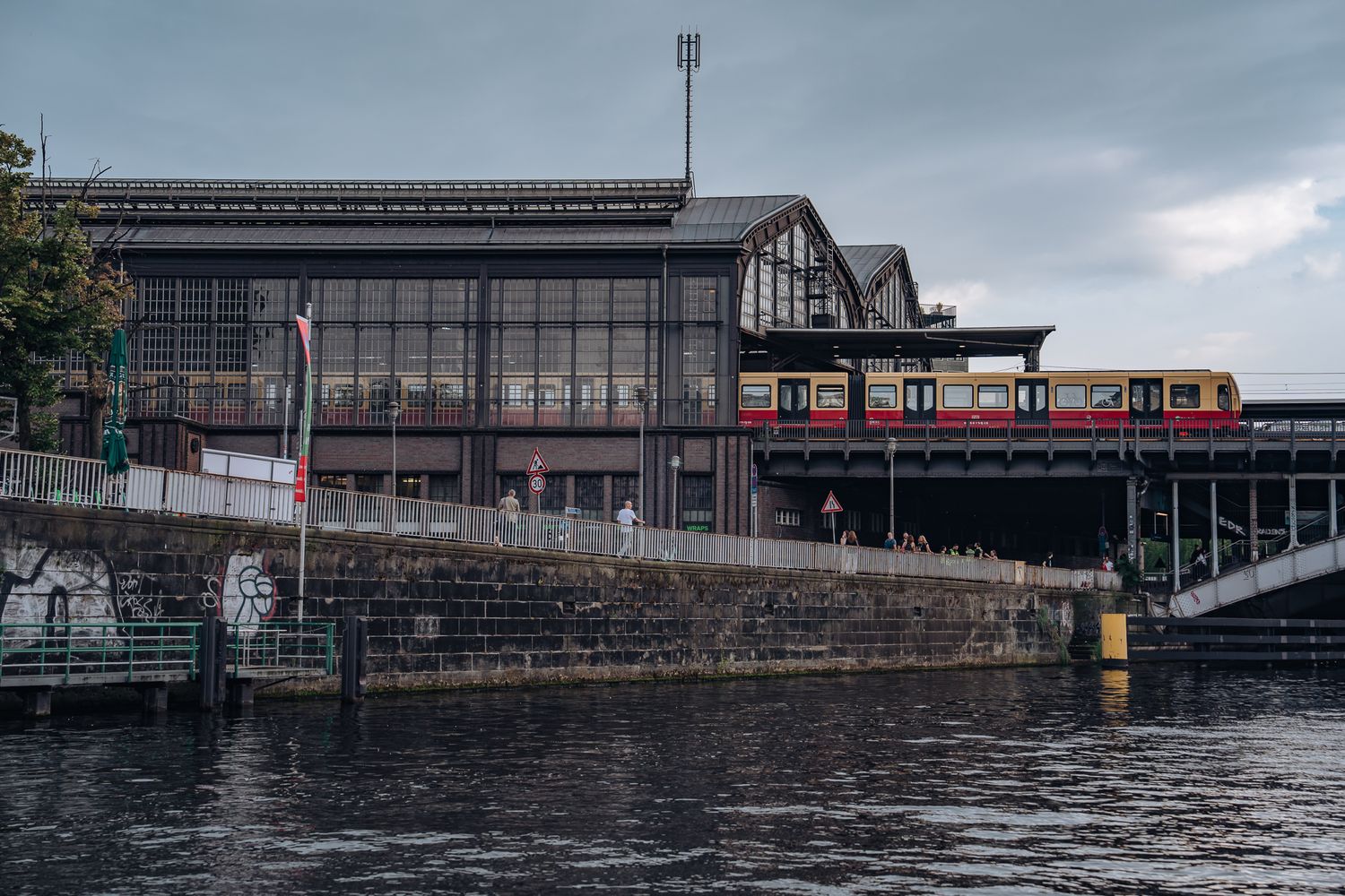 Rot-beige Berliner S-Bahn auf einem Hochgleis neben einem historischen Bahnhofgebäude mit Glasfront, davor die Spree