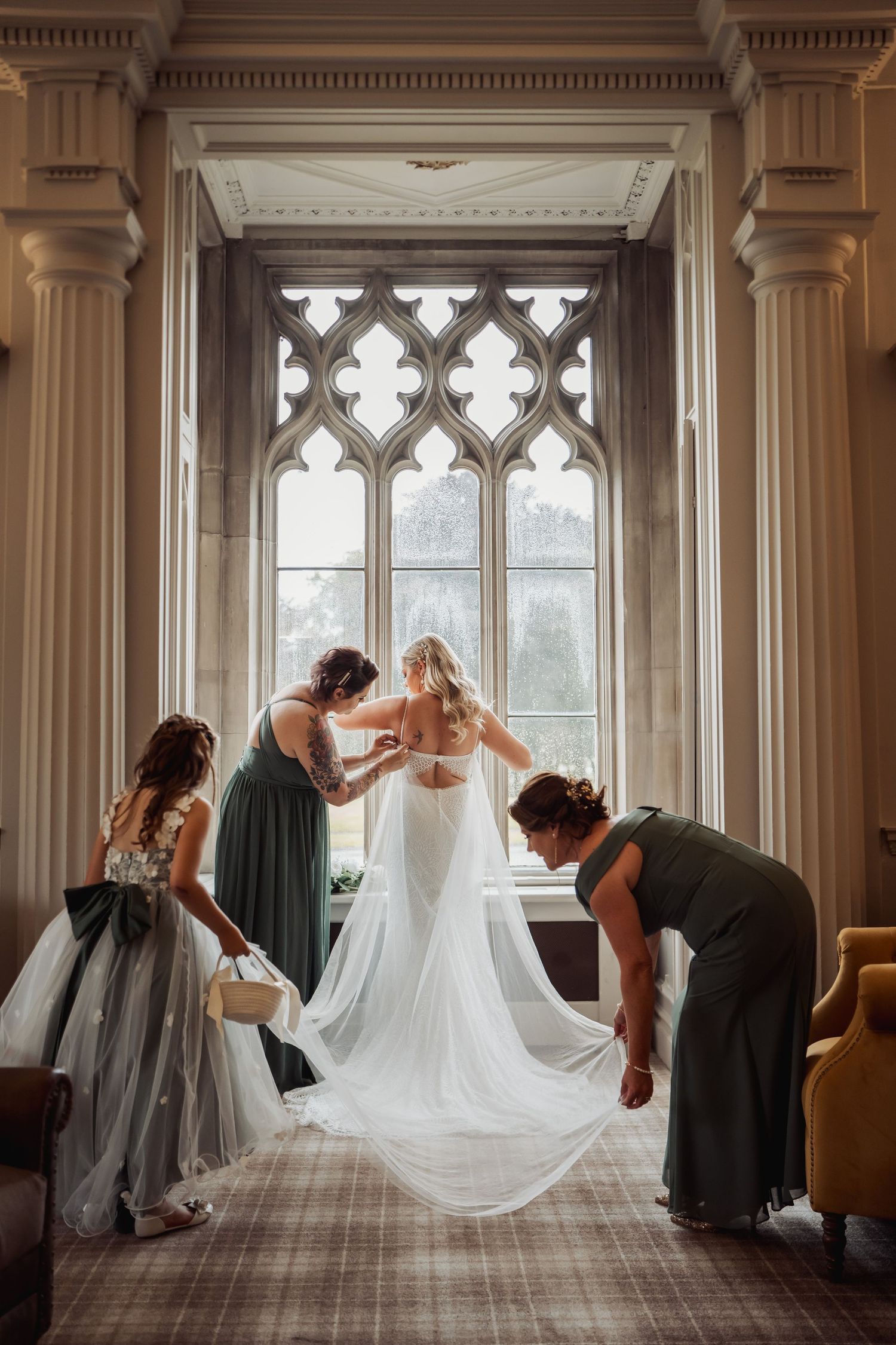 Bride being helped into her wedding dress by bridesmaids in front of a gothic arched window in an elegant venue.