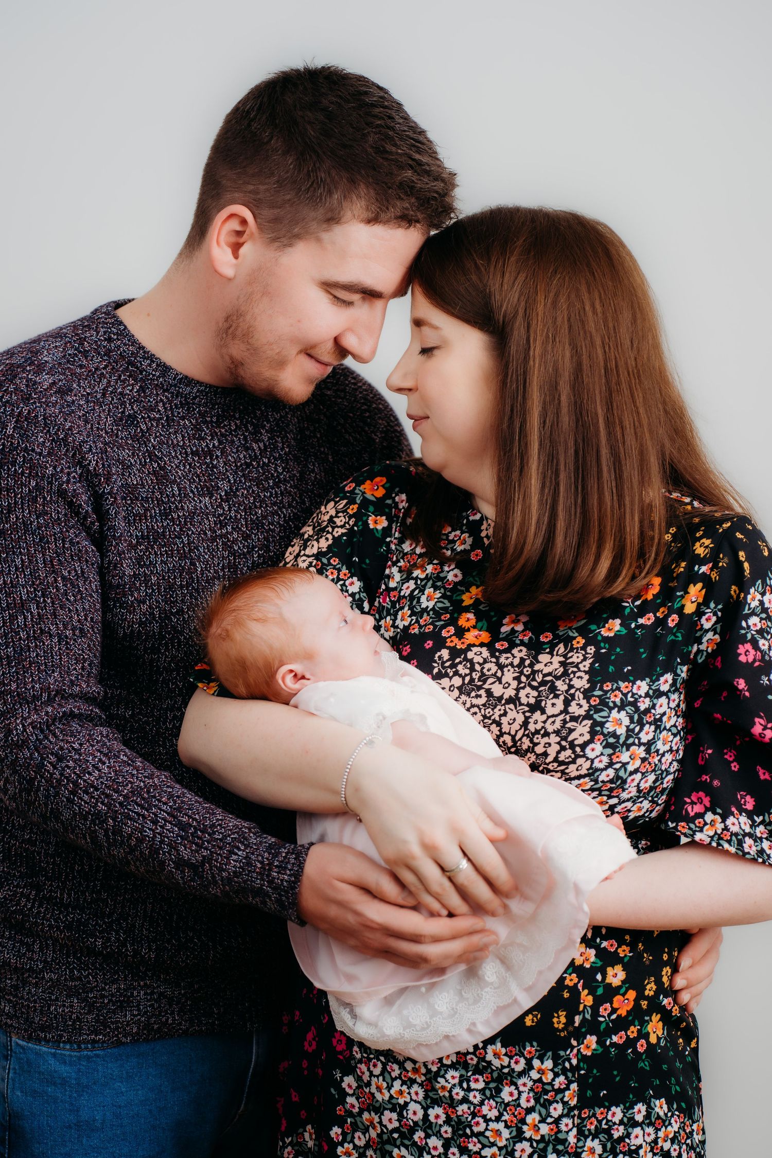 Tender family moment as parents embrace their newborn baby against gray backdrop.