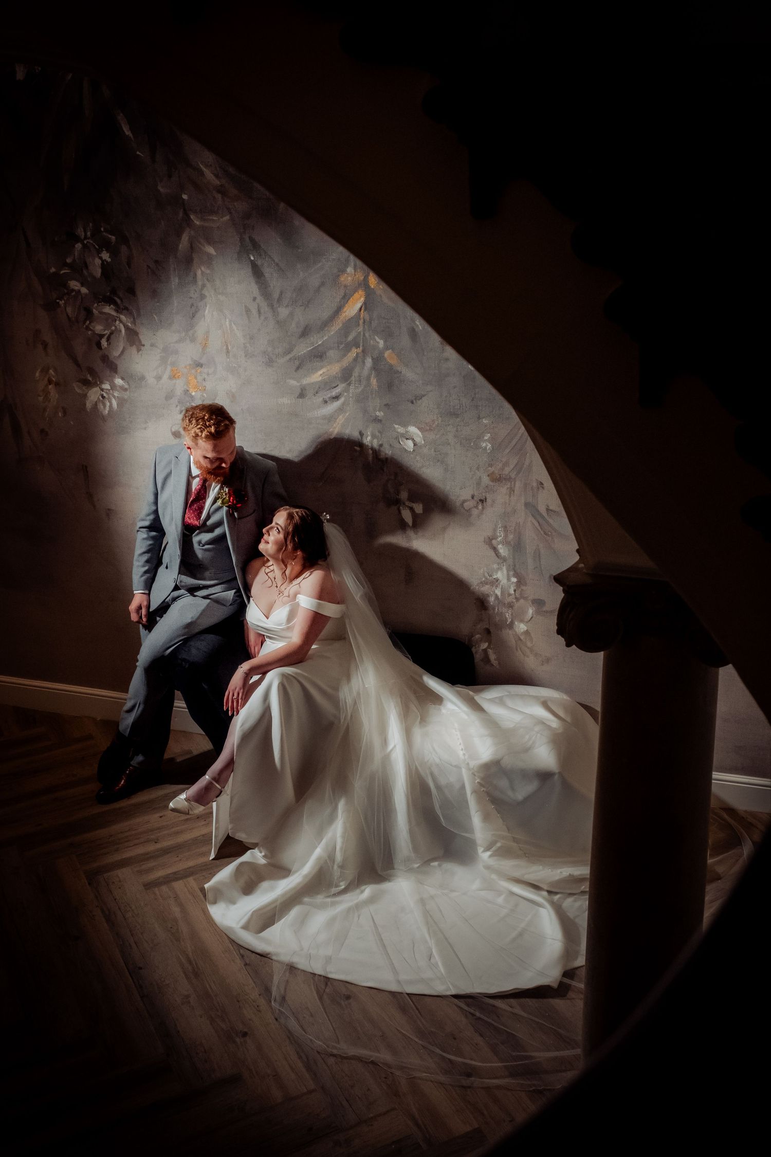 Bride in white gown and groom in blue suit share an intimate moment on a staircase in dramatic lighting.
