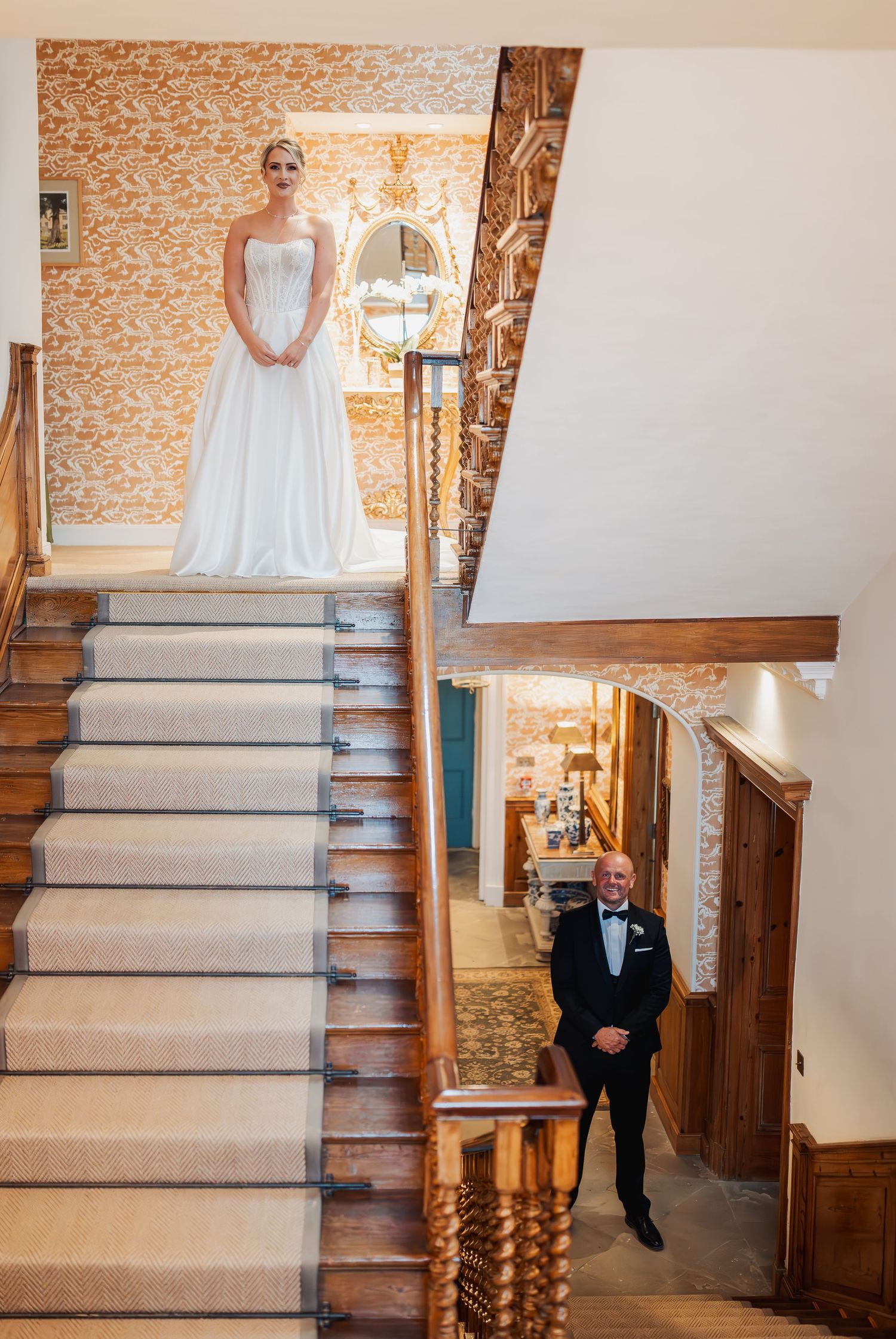 Bride in white gown stands atop grand staircase as father in tuxedo waits below in elegant venue.
