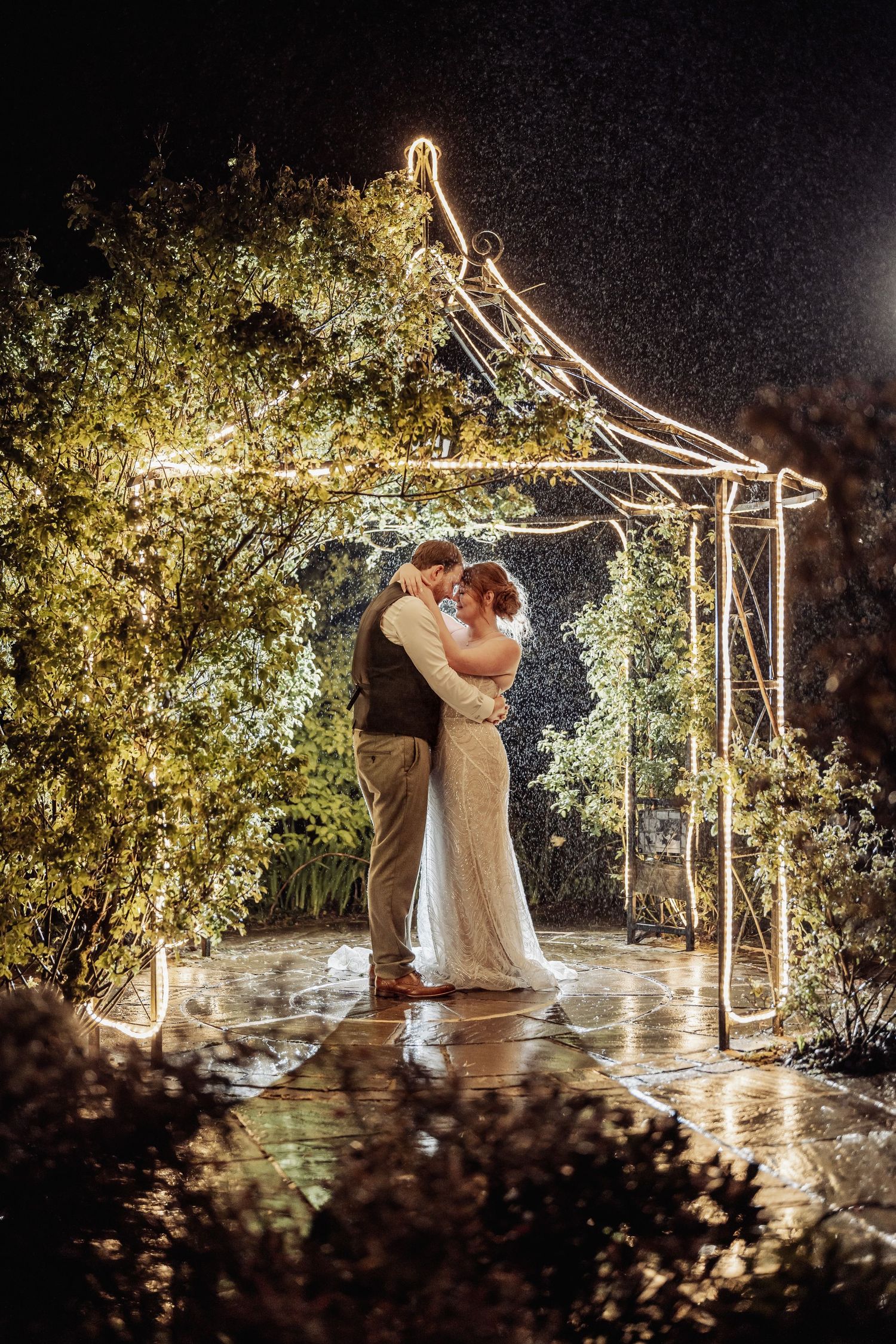 Bride and groom embrace under a glowing fairy light gazebo in the rain at night, surrounded by lush greenery.