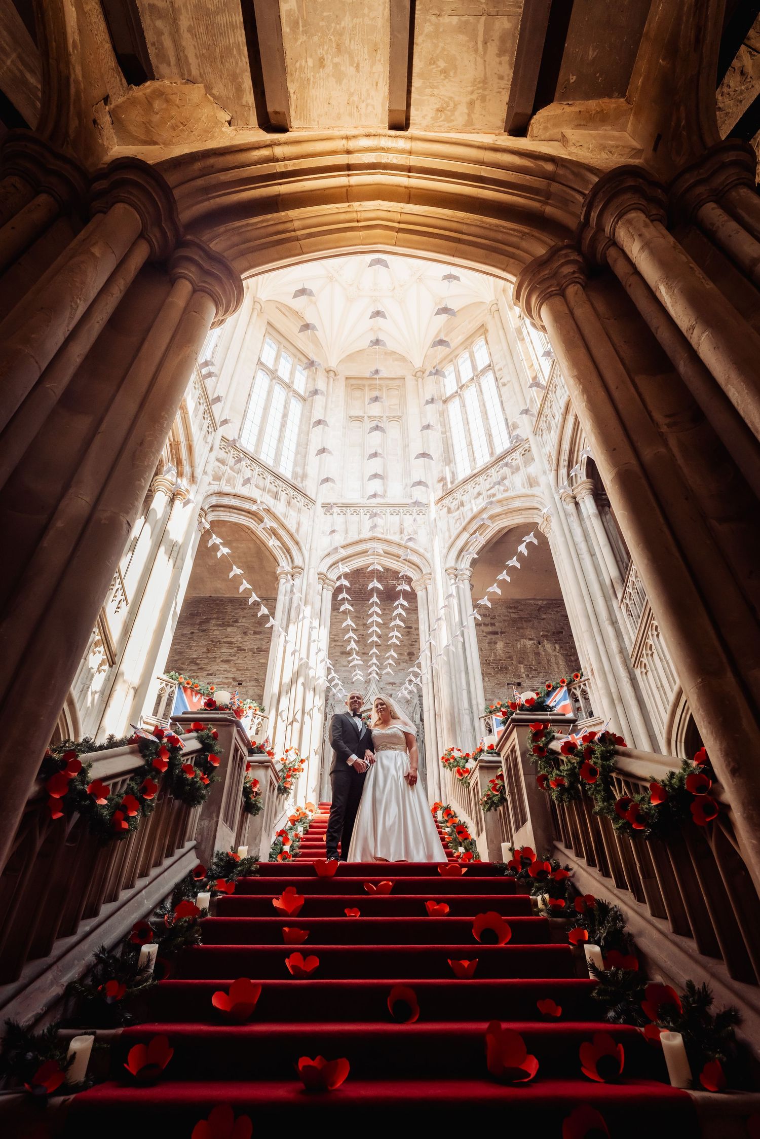 Bride and groom stand on red-carpeted stairs in a grand cathedral adorned with red roses and stunning Gothic architecture.