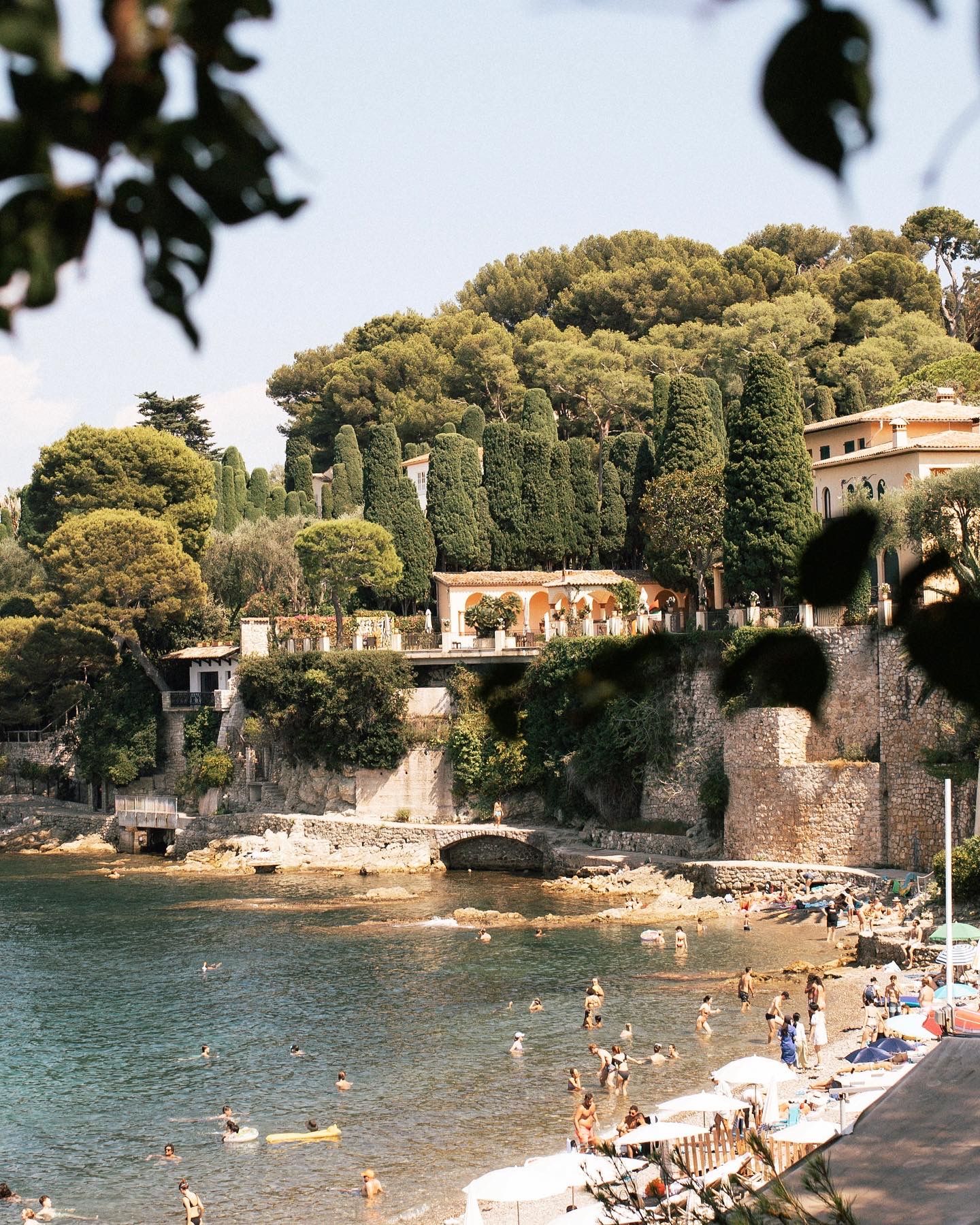 Sun-drenched Italian Riviera beach with swimmers, colorful umbrellas, stone walls, and lush green trees framing elegant hillside villas.