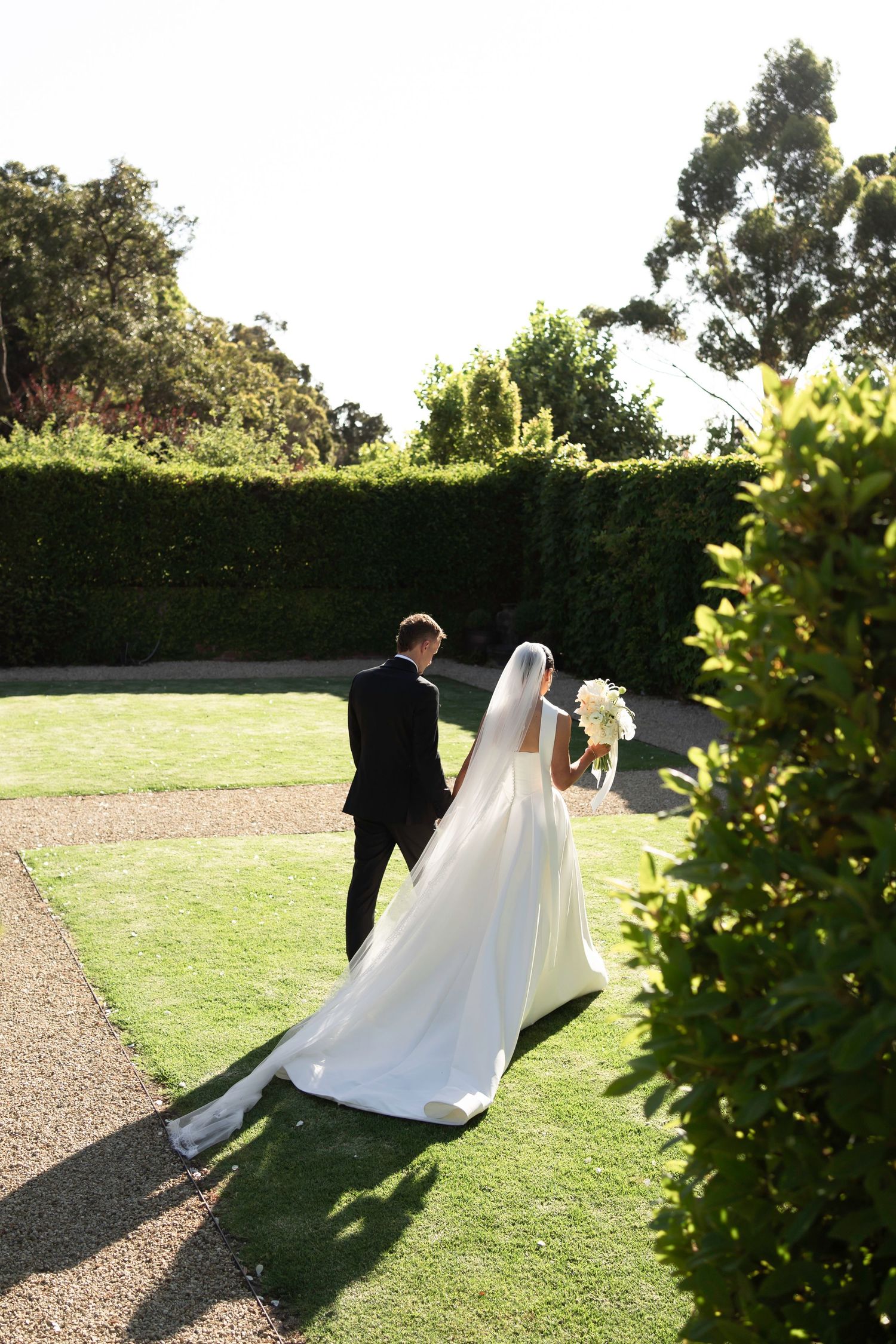 A couple in wedding attire walks through a sunlit garden path creating a romantic silhouette against the green lawn.