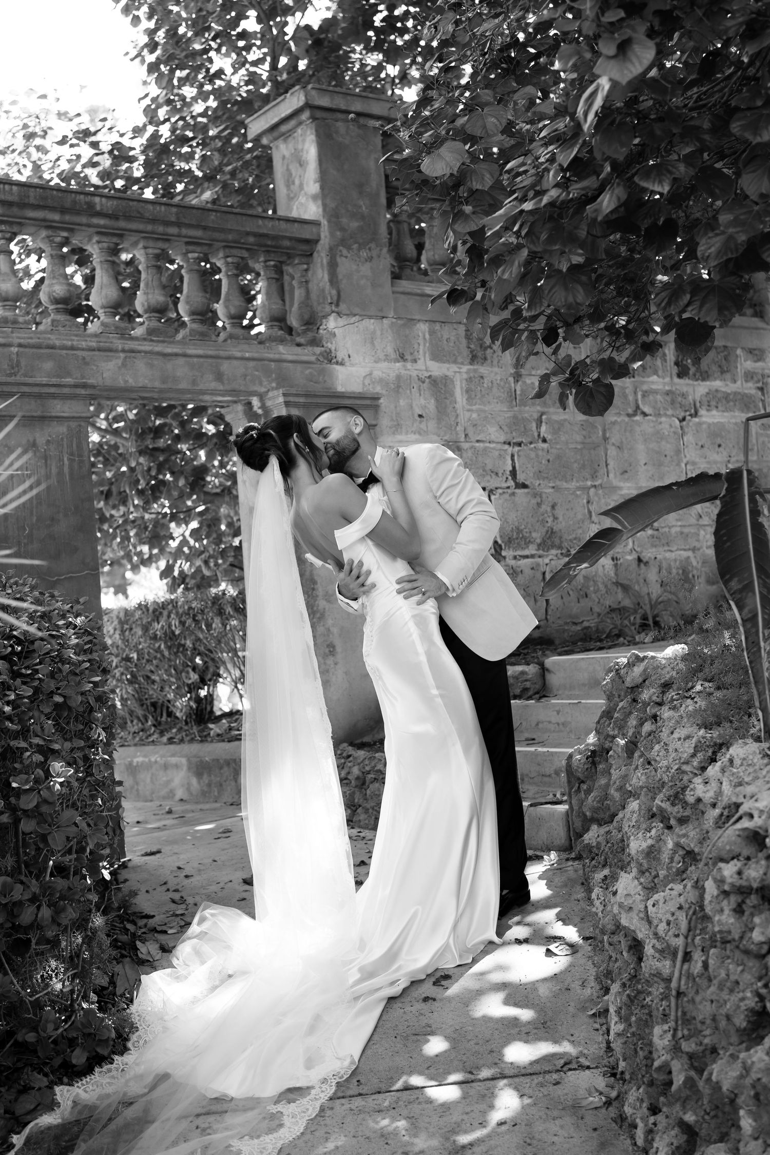 Black and white photo of a groom dipping his bride for a kiss in a romantic outdoor garden setting.
