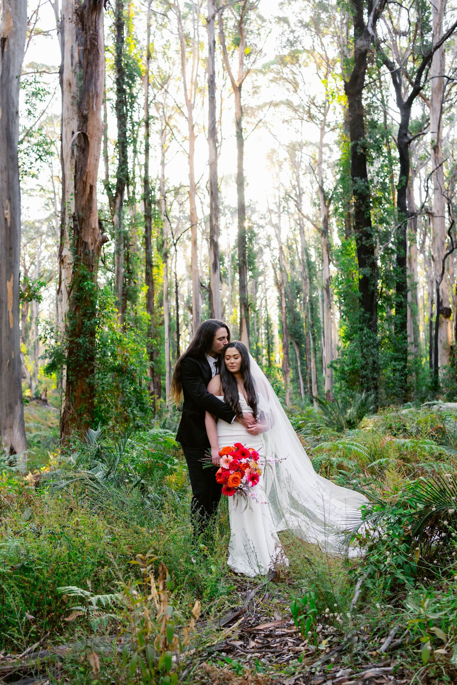 A bride in a white wedding dress poses gracefully among tall eucalyptus trees in a lush forest setting.