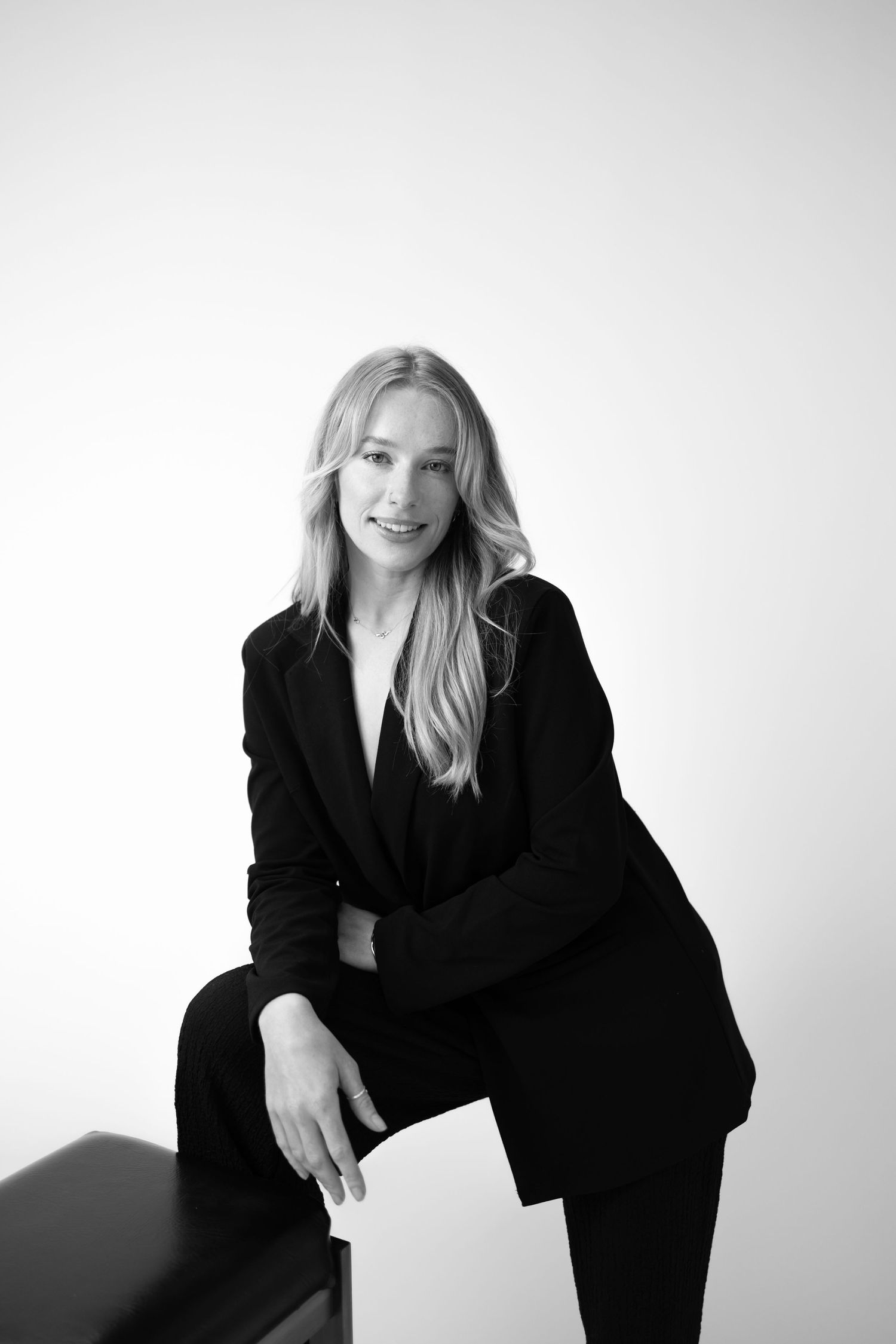 Professional black and white portrait of someone in a dark business suit sitting casually on a surface against light background.