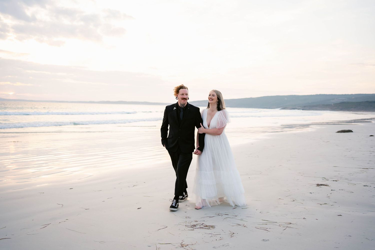 A couple walks together on a serene beach at sunset wearing formal wedding attire.