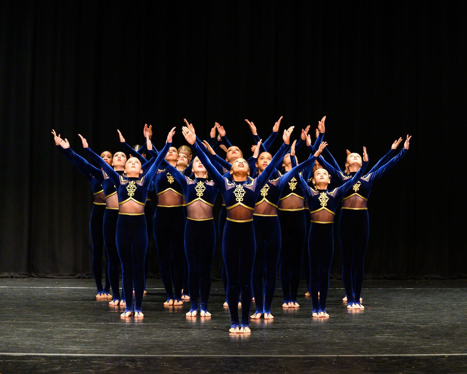 A group of dancers in navy blue uniforms perform a synchronized jump pose with their arms raised against a dark background.