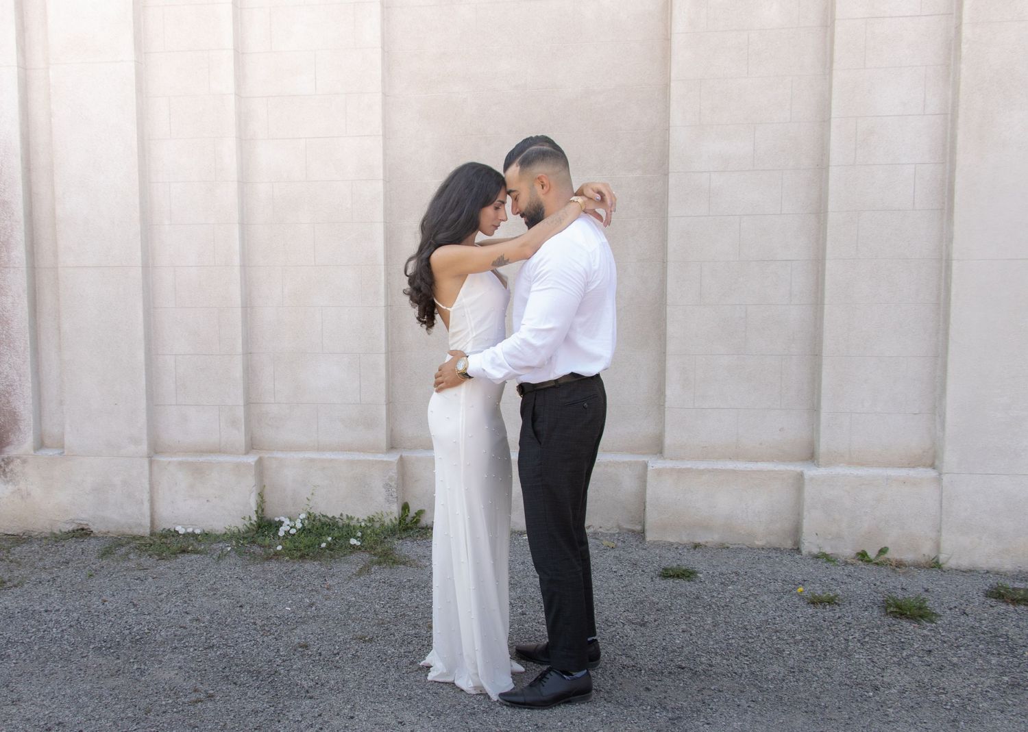 A couple stands outside in front of a beige stone wall, embracing each other. The woman, in a long white dress, and the 