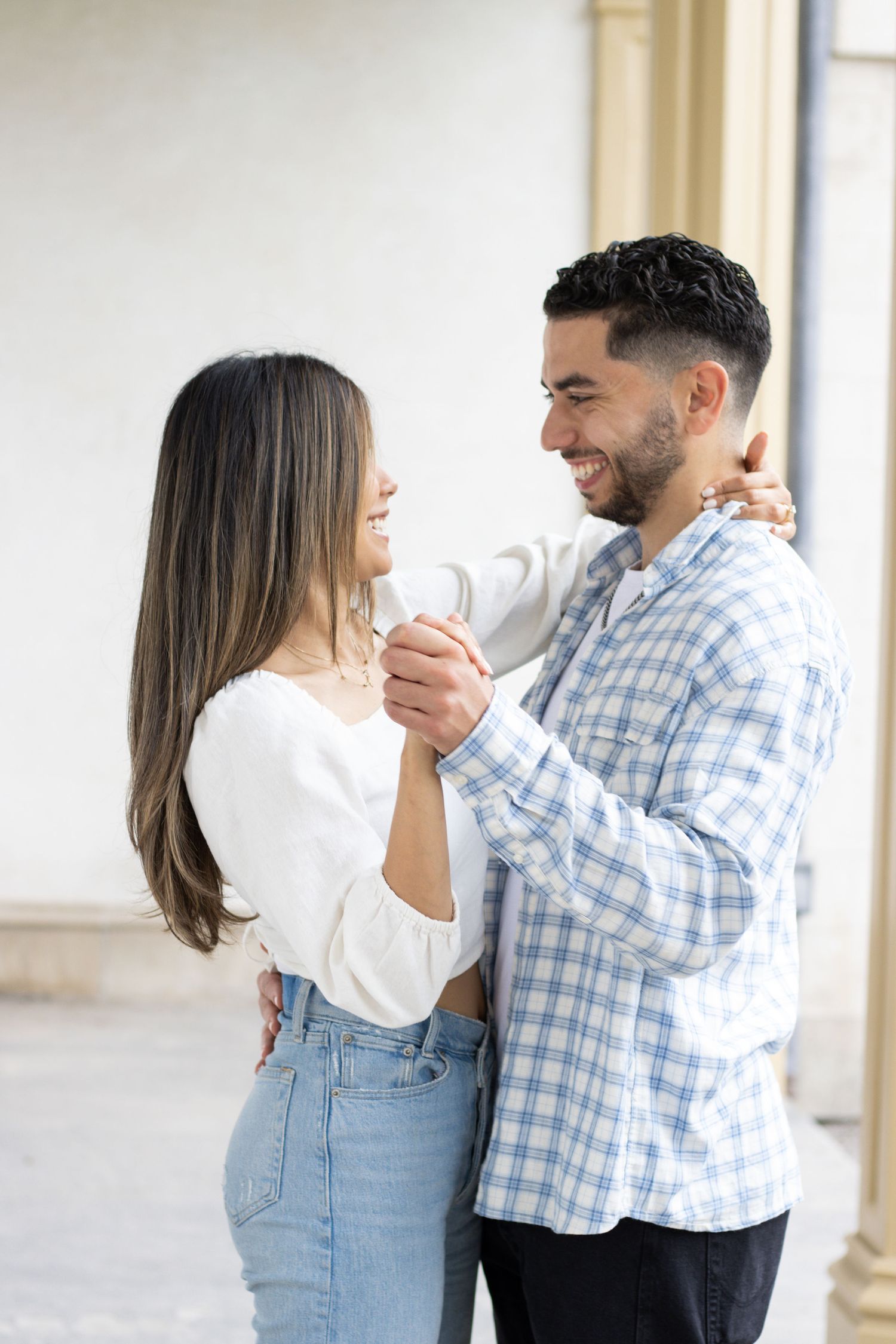 A couple slow dancing, in an outdoor corridor. The woman is wearing a white long-sleeve top and blue jeans, while the ma