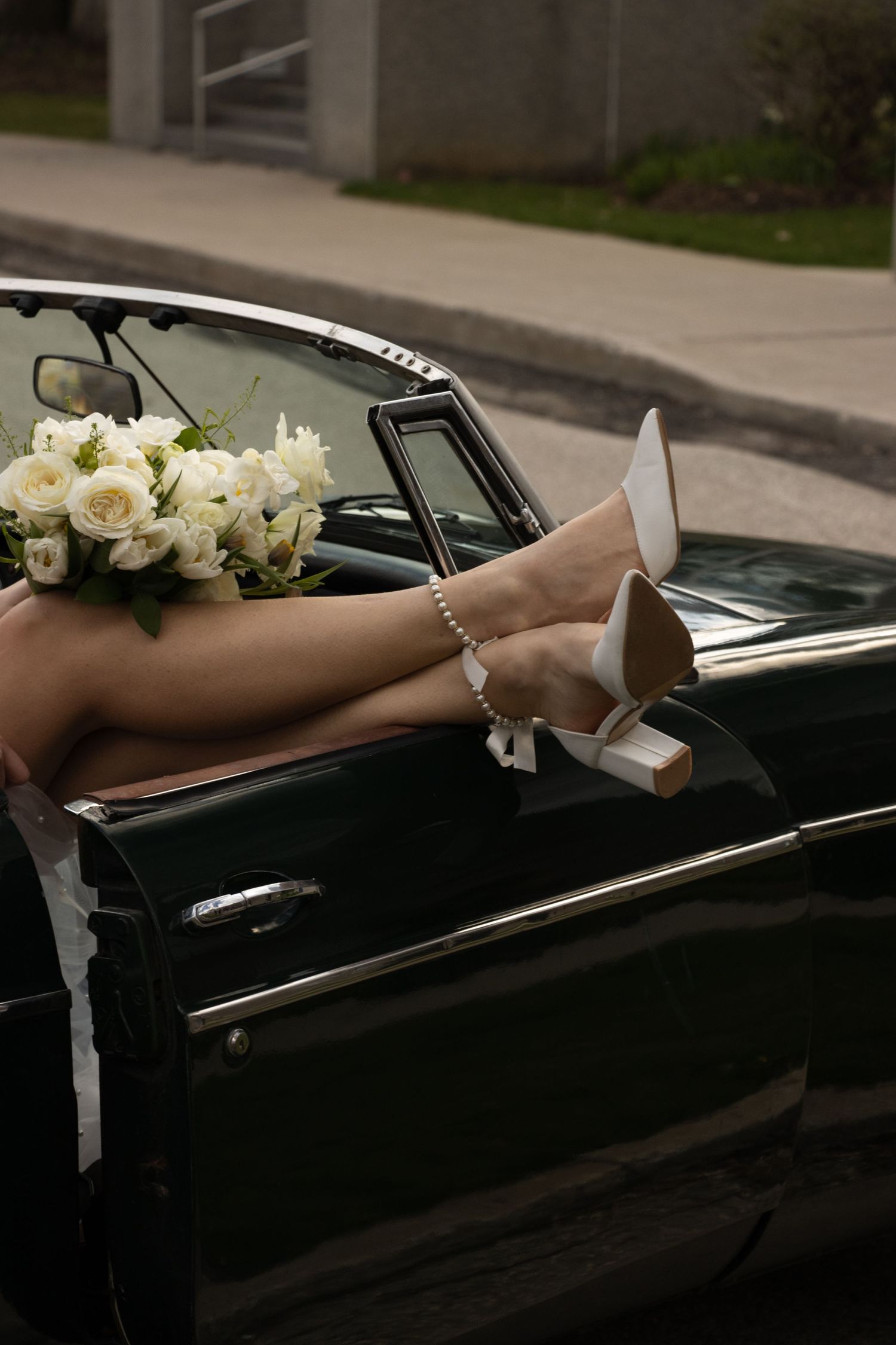 A pair of legs in white high heels is seen resting on the door of a vintage green convertible car. The person holds a bo
