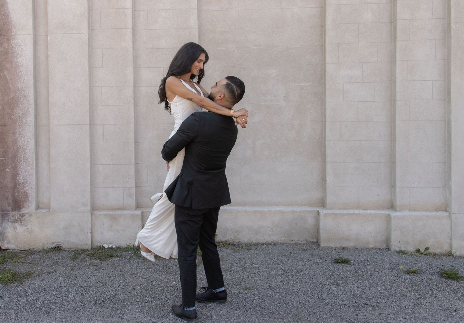 A woman, dressed in a white dress and heels, is being lifted off the ground by a man wearing a black suit. They are posi