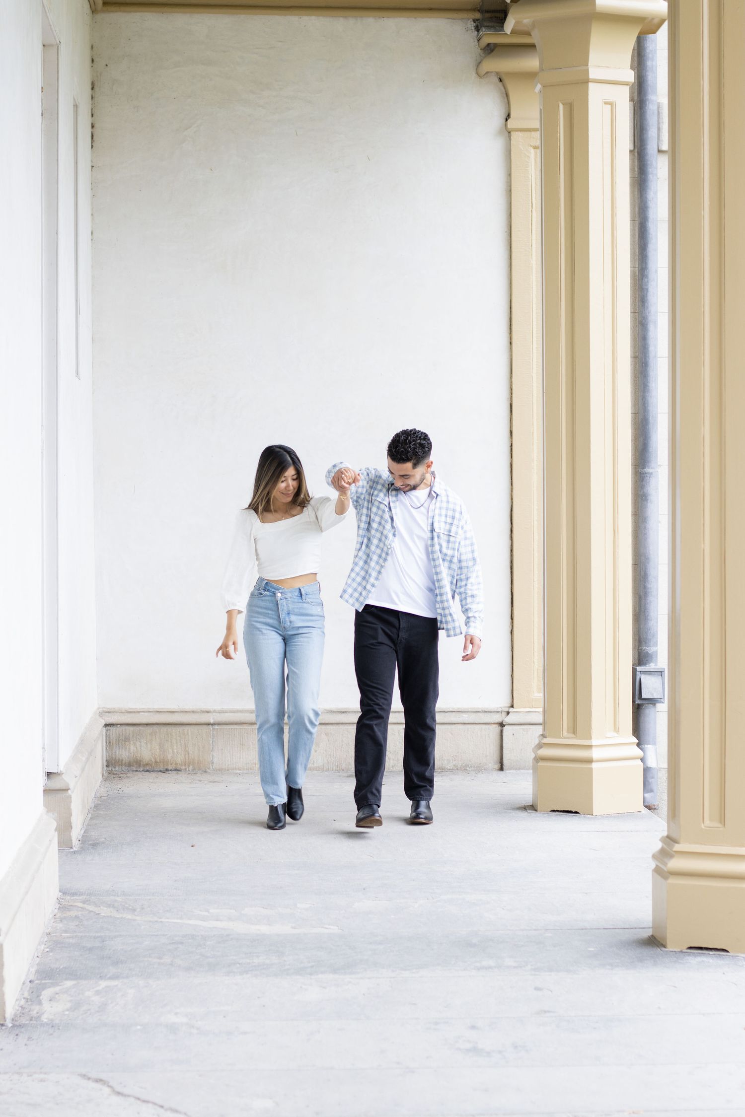 A couple holding hands walks together in an outdoor corridor. The woman is wearing a white long-sleeve top and blue jean