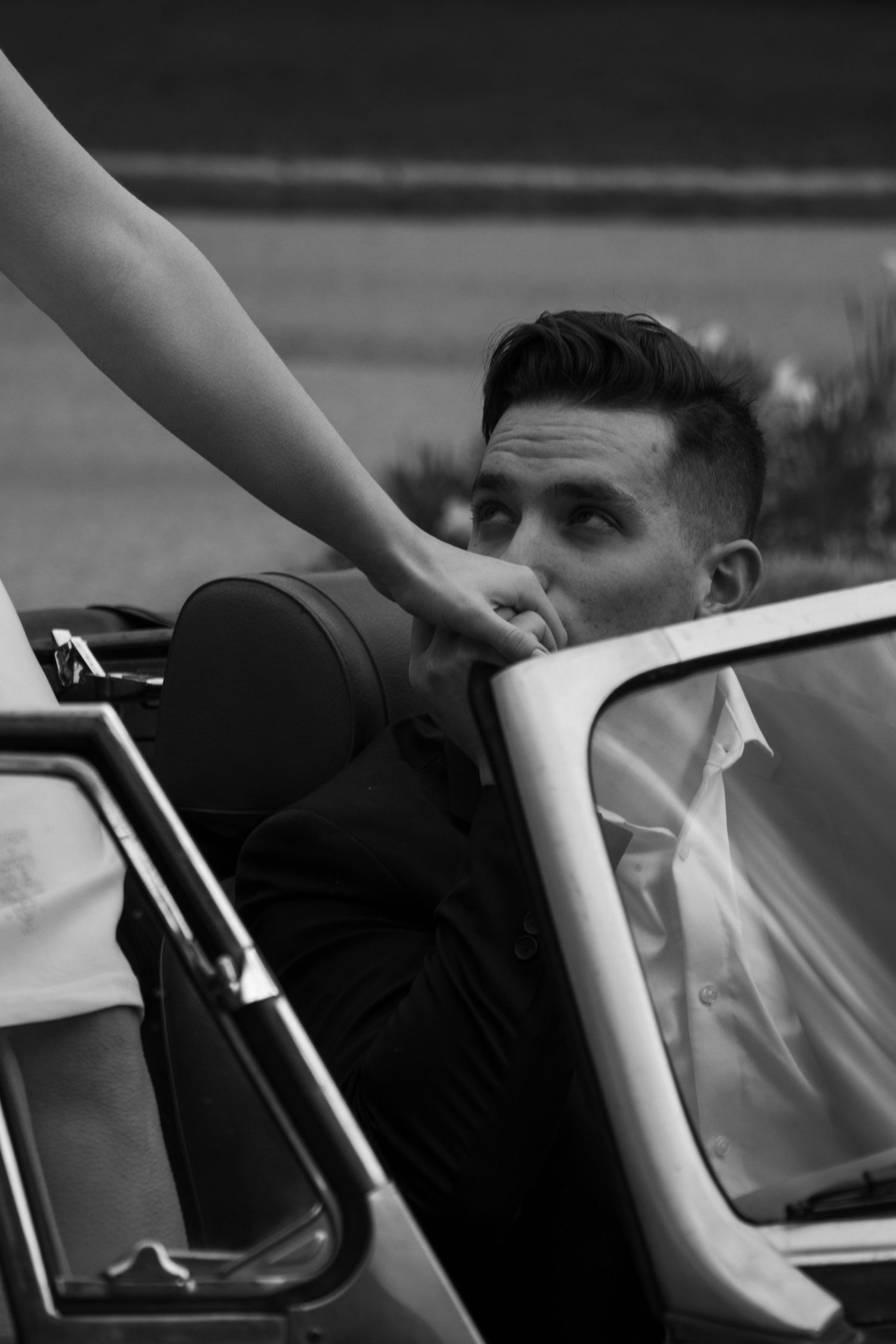 A well-dressed man in a suit is seated in a classic convertible car, gently kissing the hand of a woman standing beside 