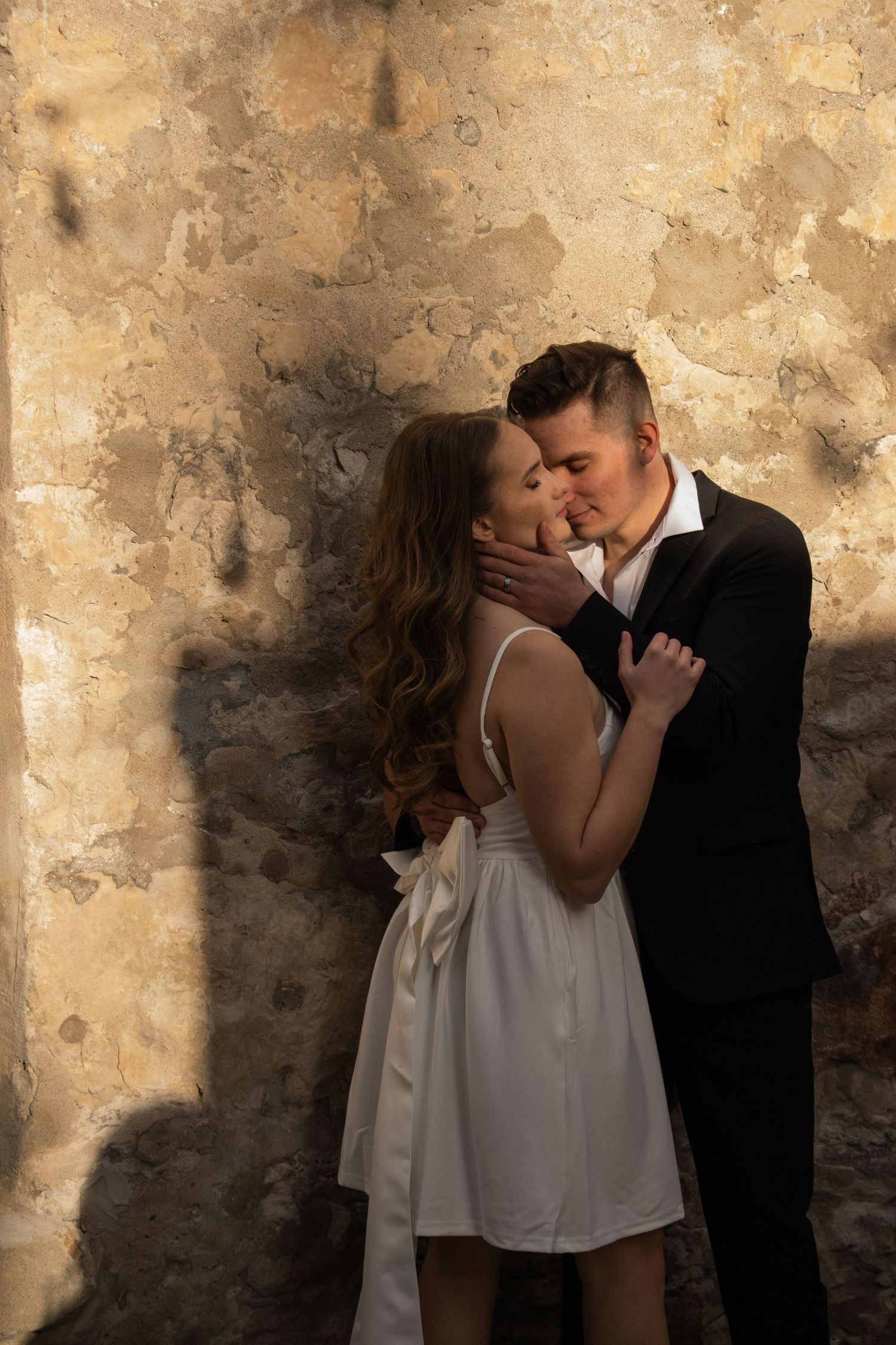 A couple stands in an intimate embrace against a rustic stone wall. The woman, wearing a white dress with a ribbon tie a