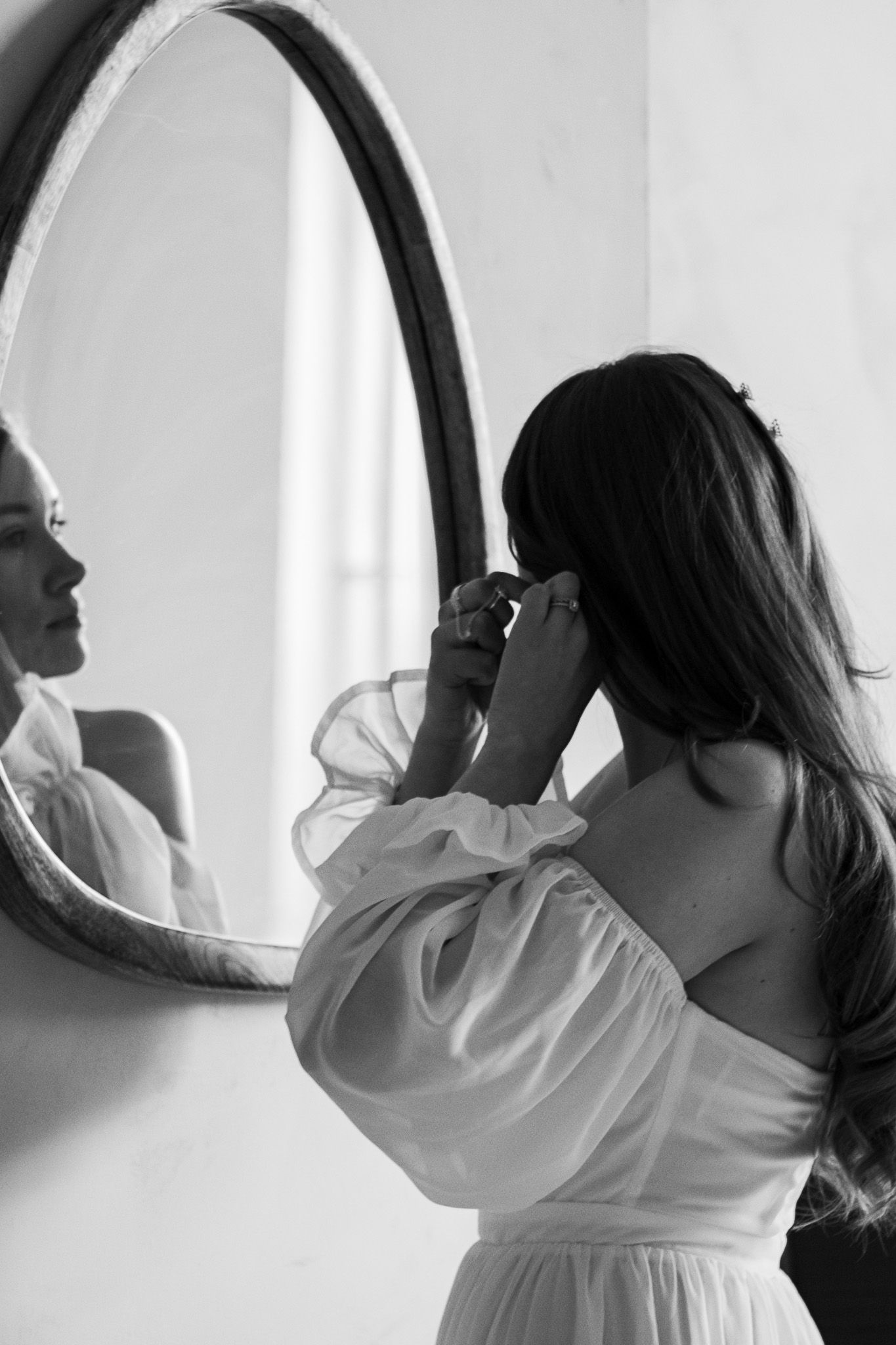 A woman with long hair stands facing a wall-mounted oval mirror, adjusting her earring. She is wearing an off-the-should