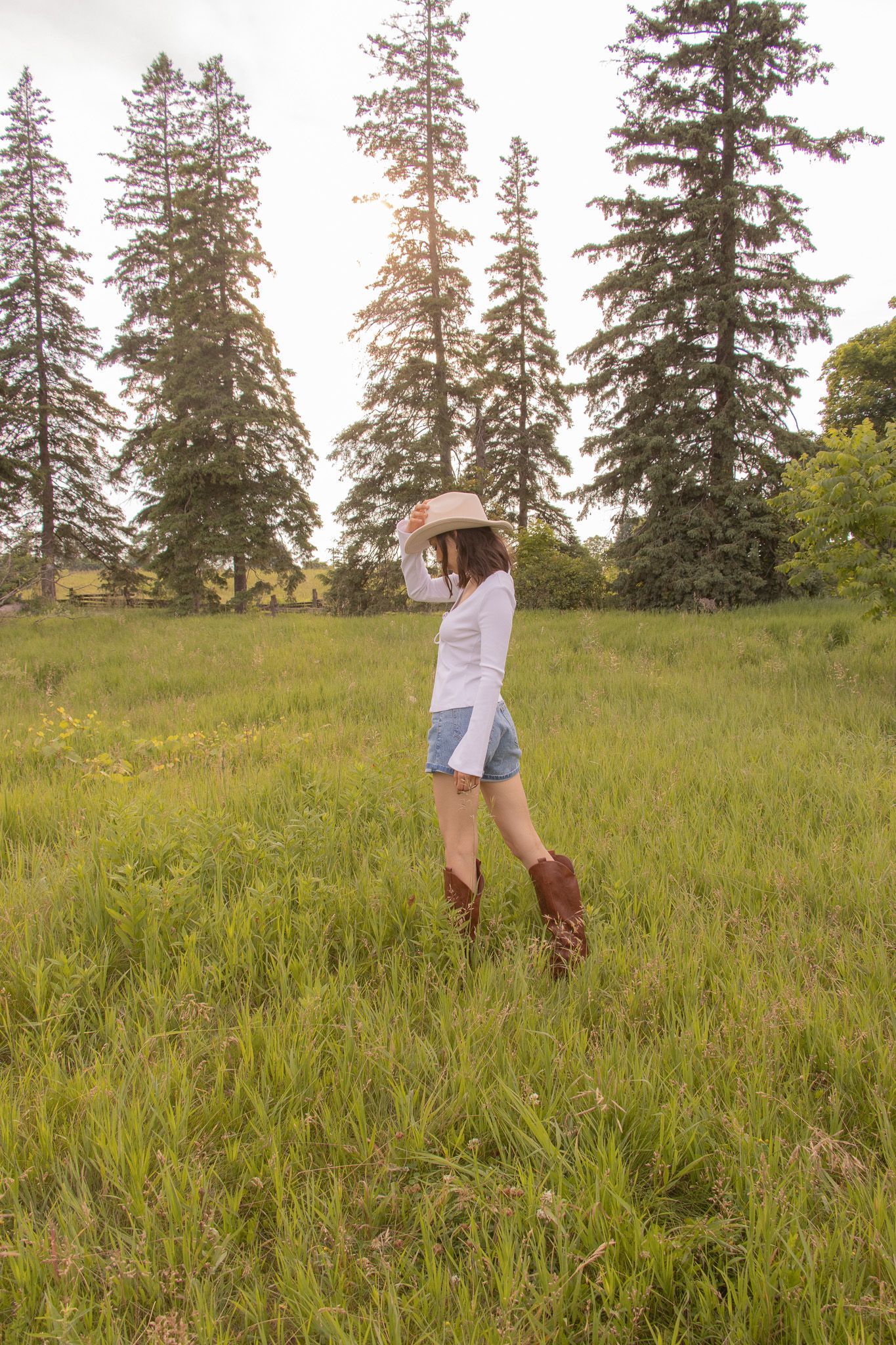 A person wearing a white long-sleeve shirt, denim shorts, brown knee-high boots, and a beige hat stands in a grassy fiel