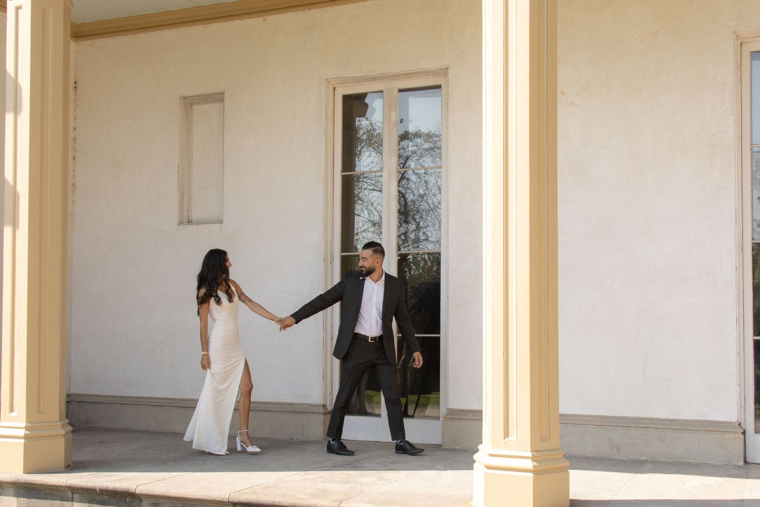 A couple, dressed elegantly, stand hand in hand on a porch with tall columns and decorative molding.
