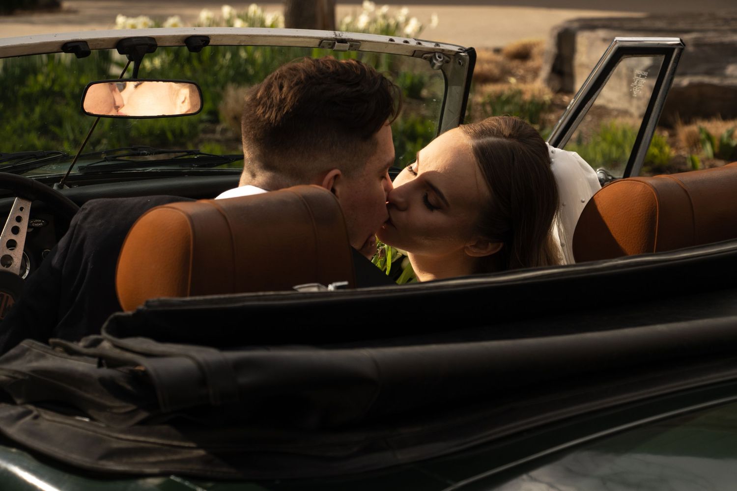 A couple is seen from behind, sitting in a dark green convertible car, sharing a kiss. The woman is wearing a wedding dr