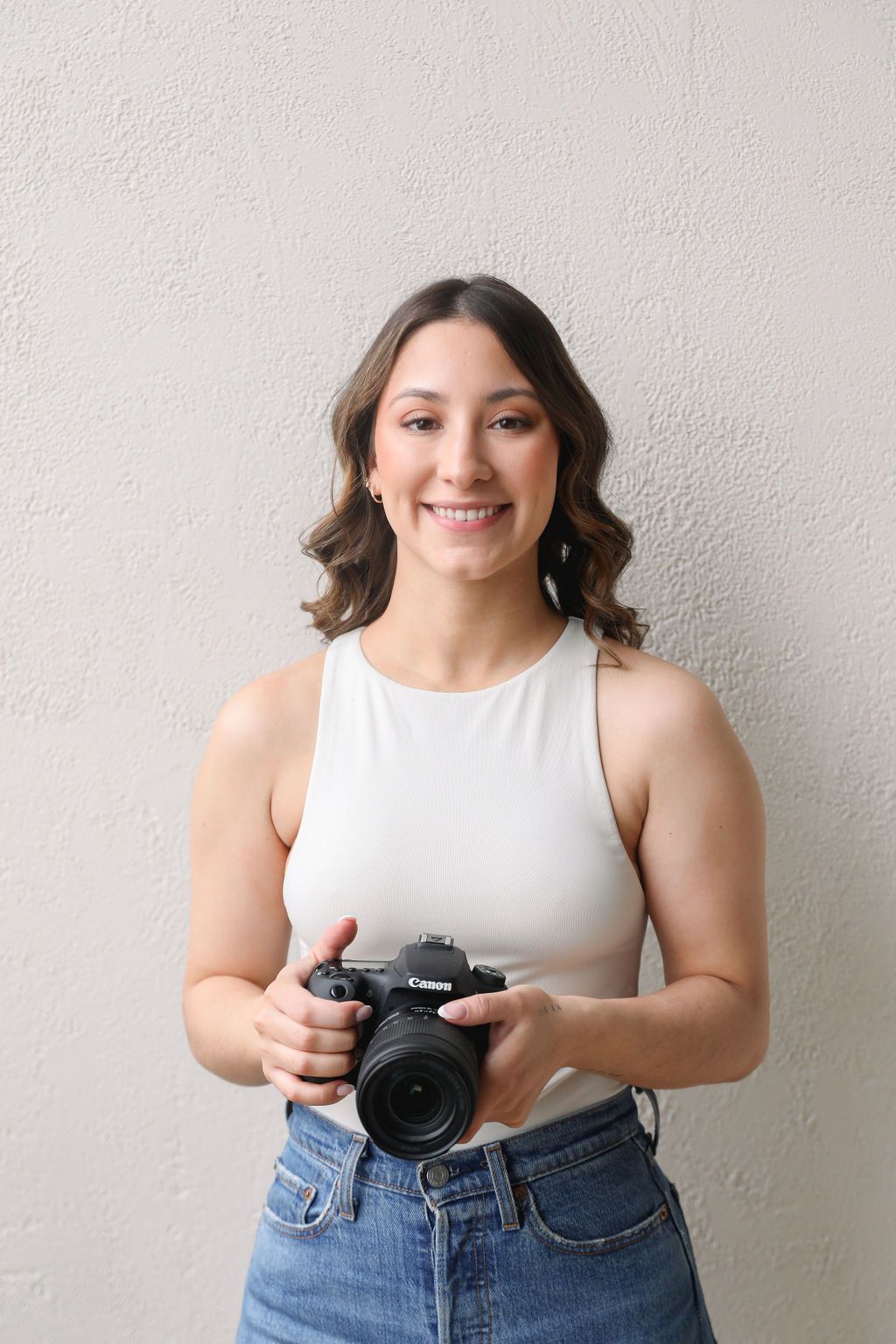 A woman with wavy brown hair, wearing a white sleeveless top and blue jeans, smiles while holding a Canon camera. She st