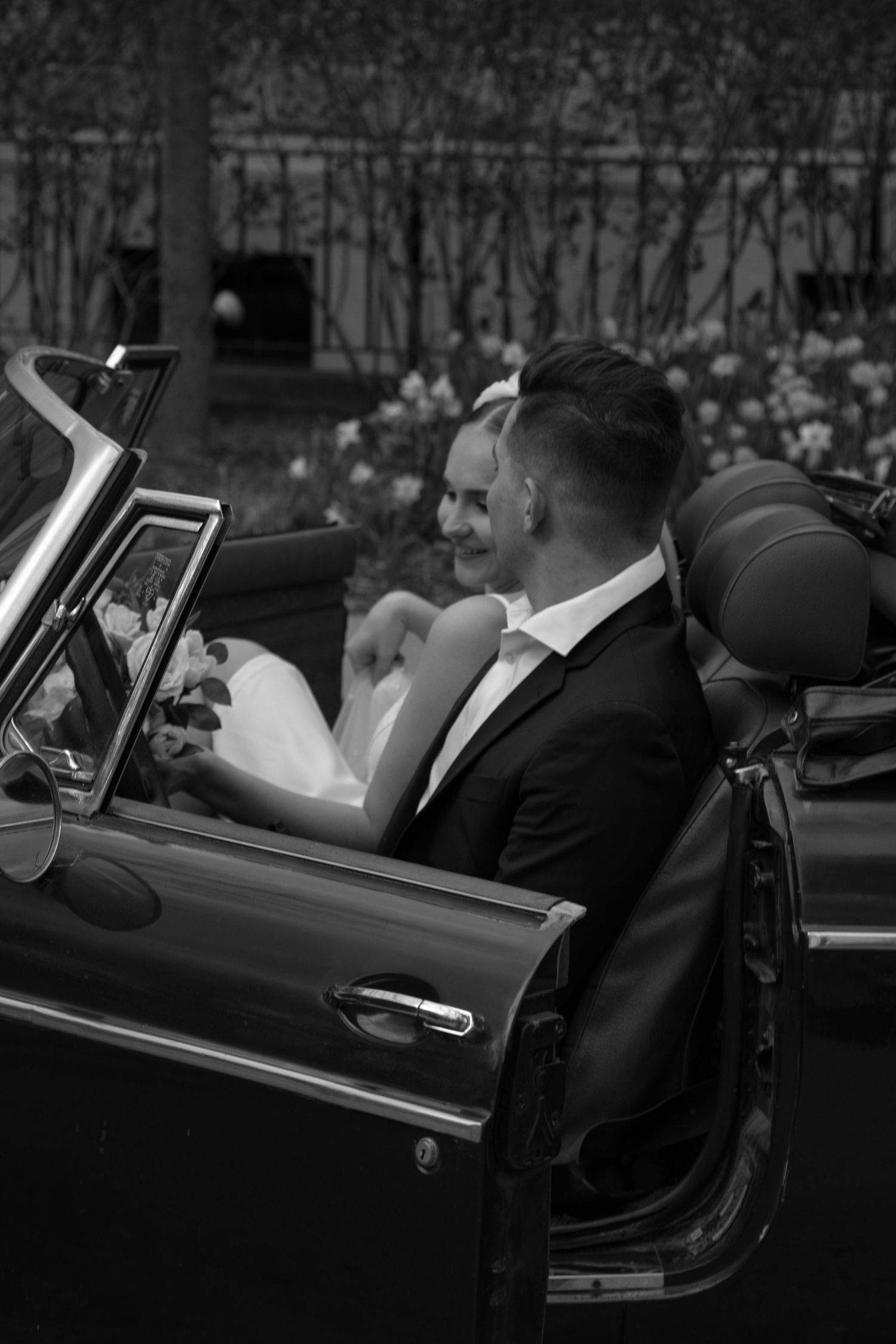A black-and-white photo captures a couple sitting in a convertible car. The woman is holding a bouquet of flowers and lo