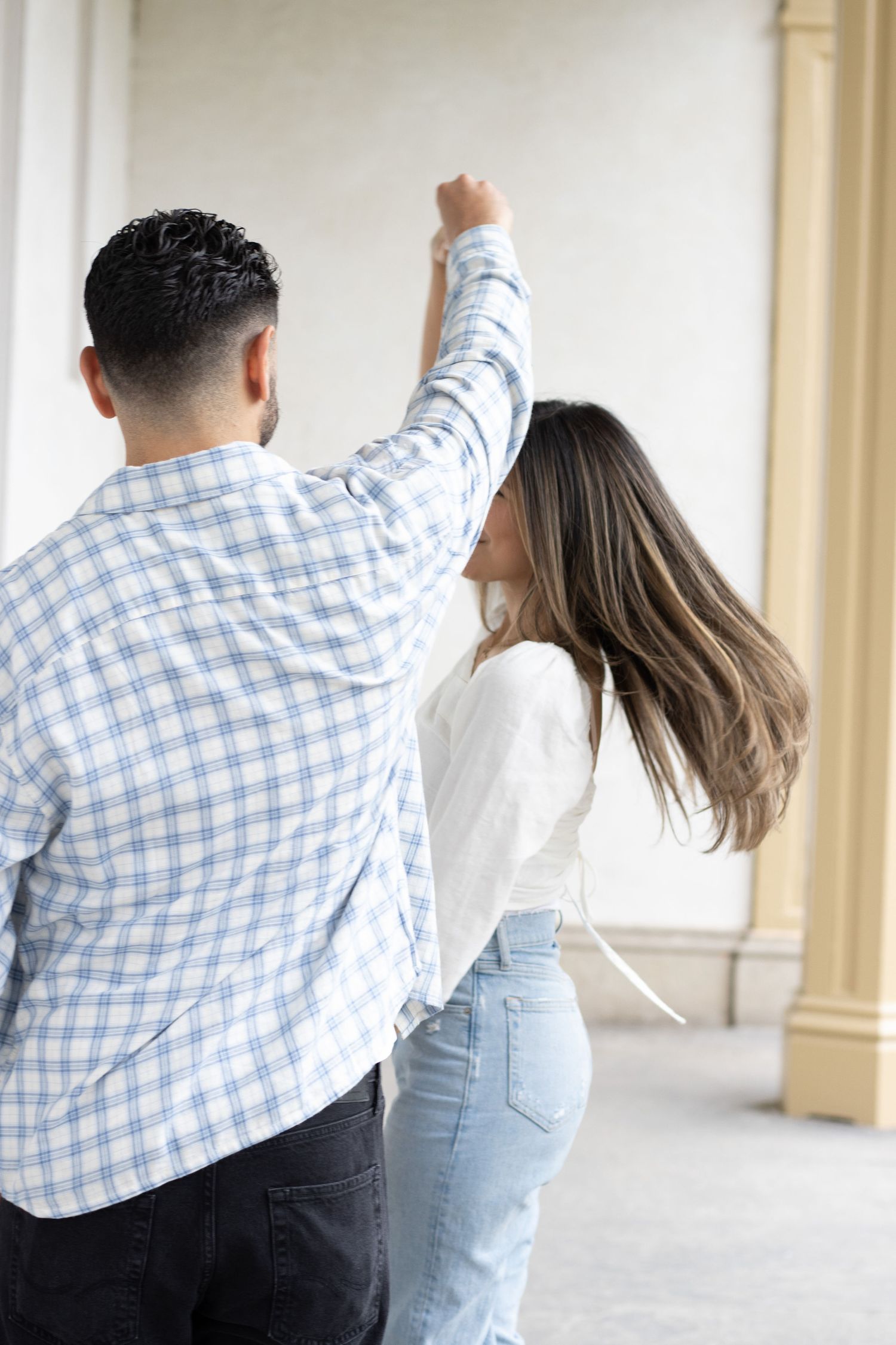A couple slow dancing, in an outdoor corridor. The man is spinning the woman. The woman is wearing a white long-sleeve t