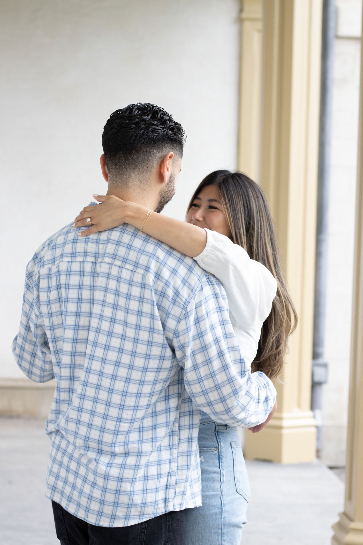 A couple slow dancing, in an outdoor corridor. The woman is wearing a white long-sleeve top and blue jeans, while the ma