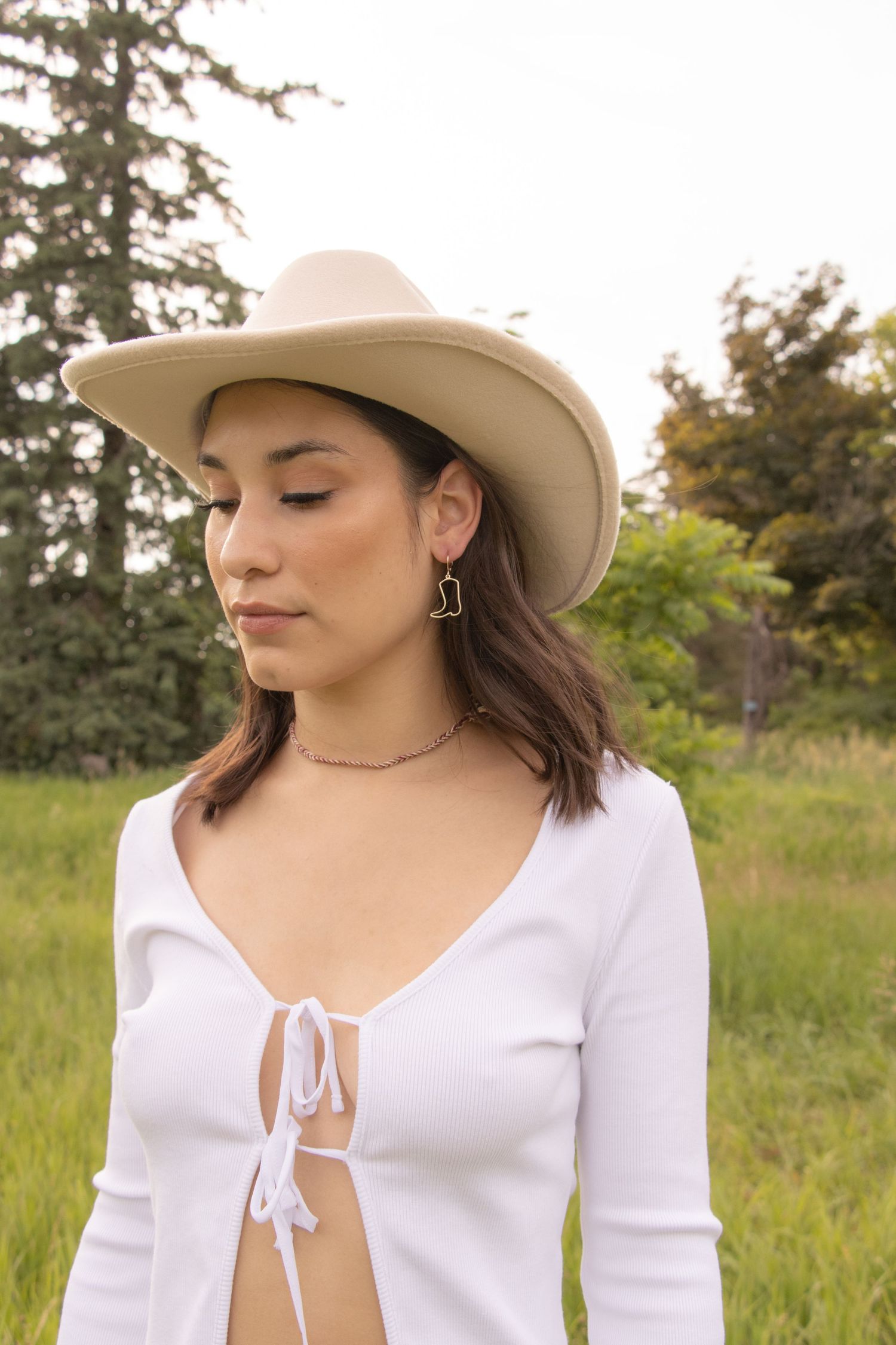 A person with shoulder-length dark hair stands in a grassy field wearing a beige cowboy hat, a white long-sleeved top wi