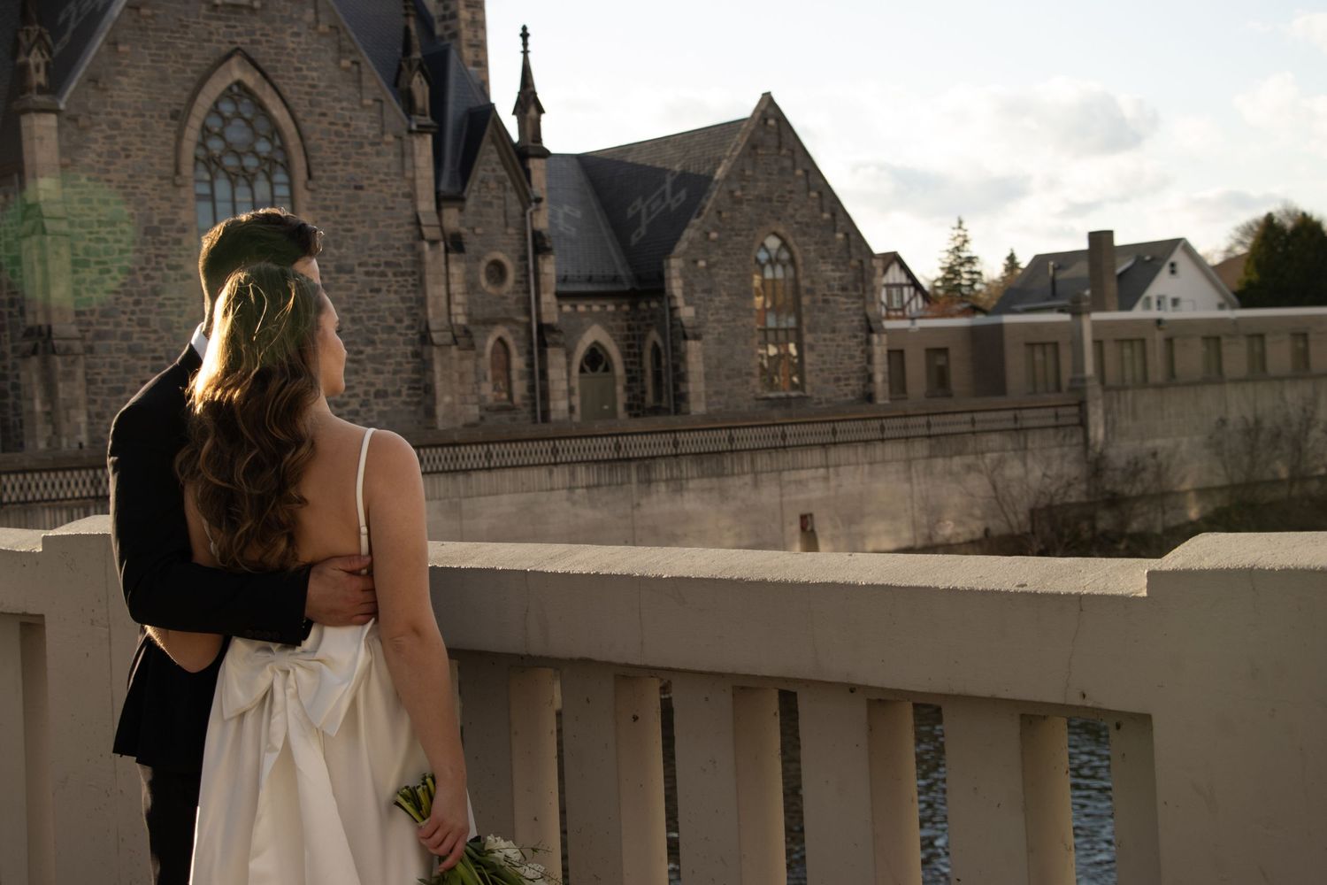 A bride and groom stand on a bridge, with the groom embracing the bride from behind. The bride holds a bouquet of white 