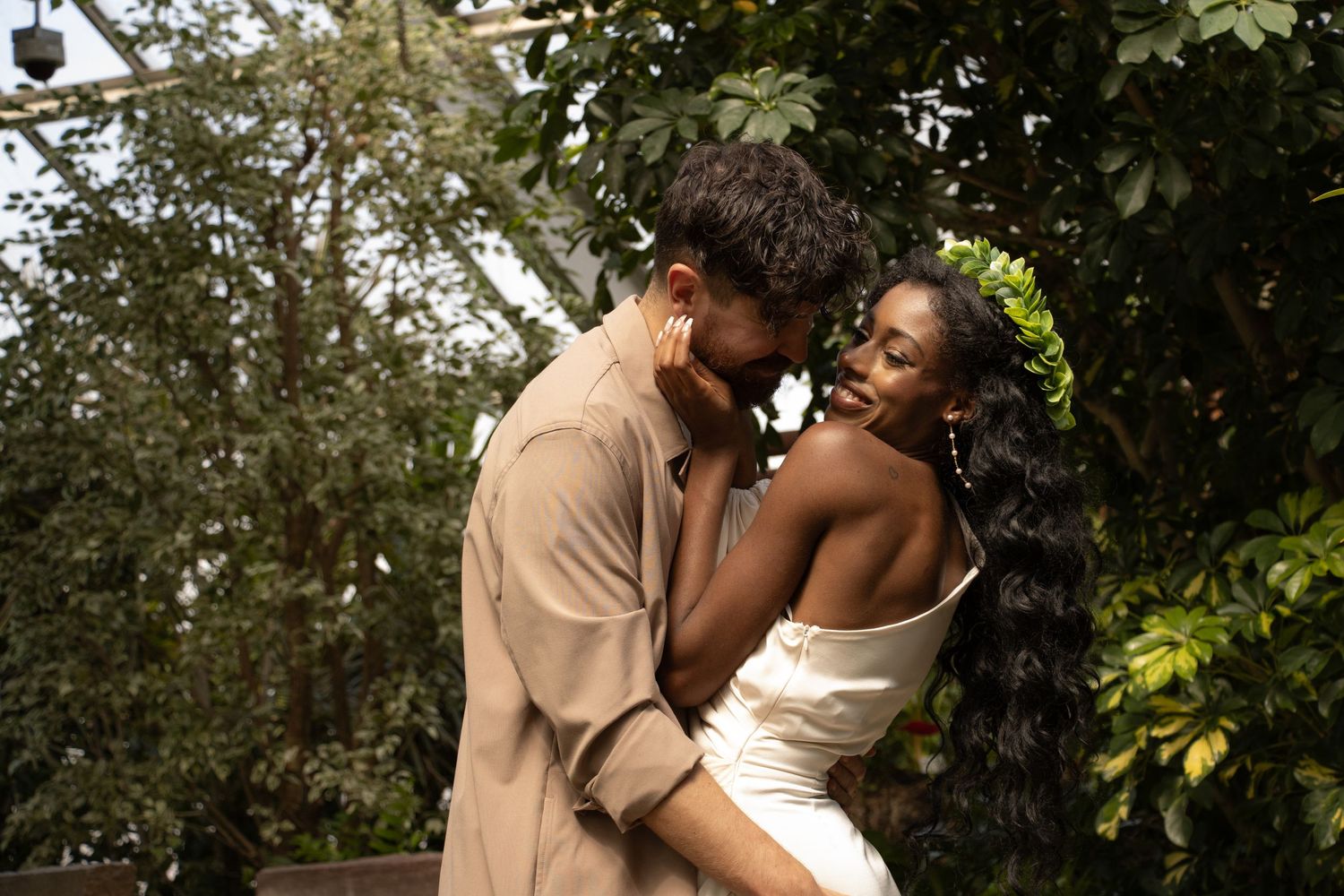 A joyful couple embraces outdoors, surrounded by lush greenery. The woman, wearing a white dress and a leafy crown, smil