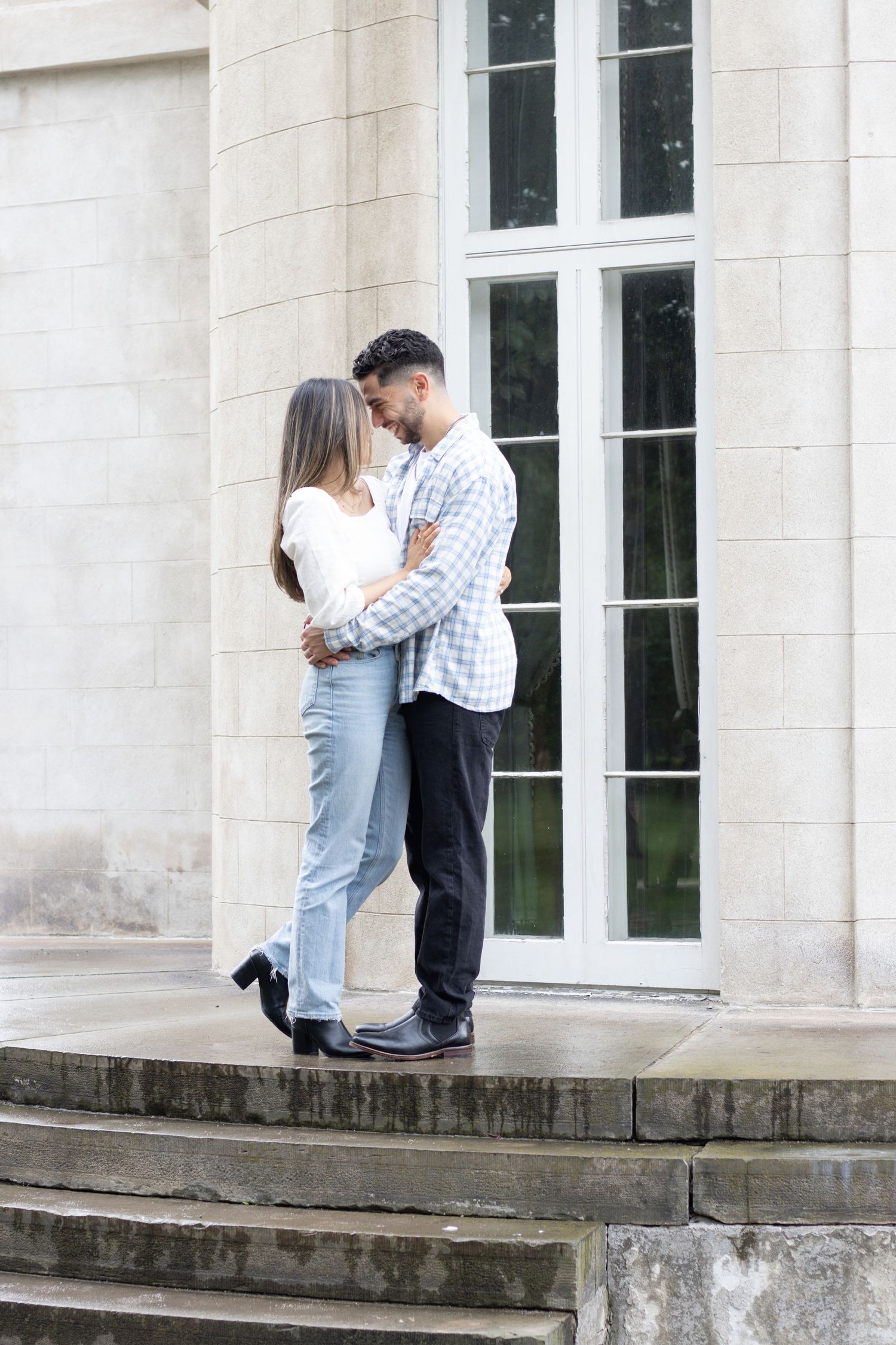 A couple stands outside a building with large windows. They are facing each other, smiling, and embracing. The woman has