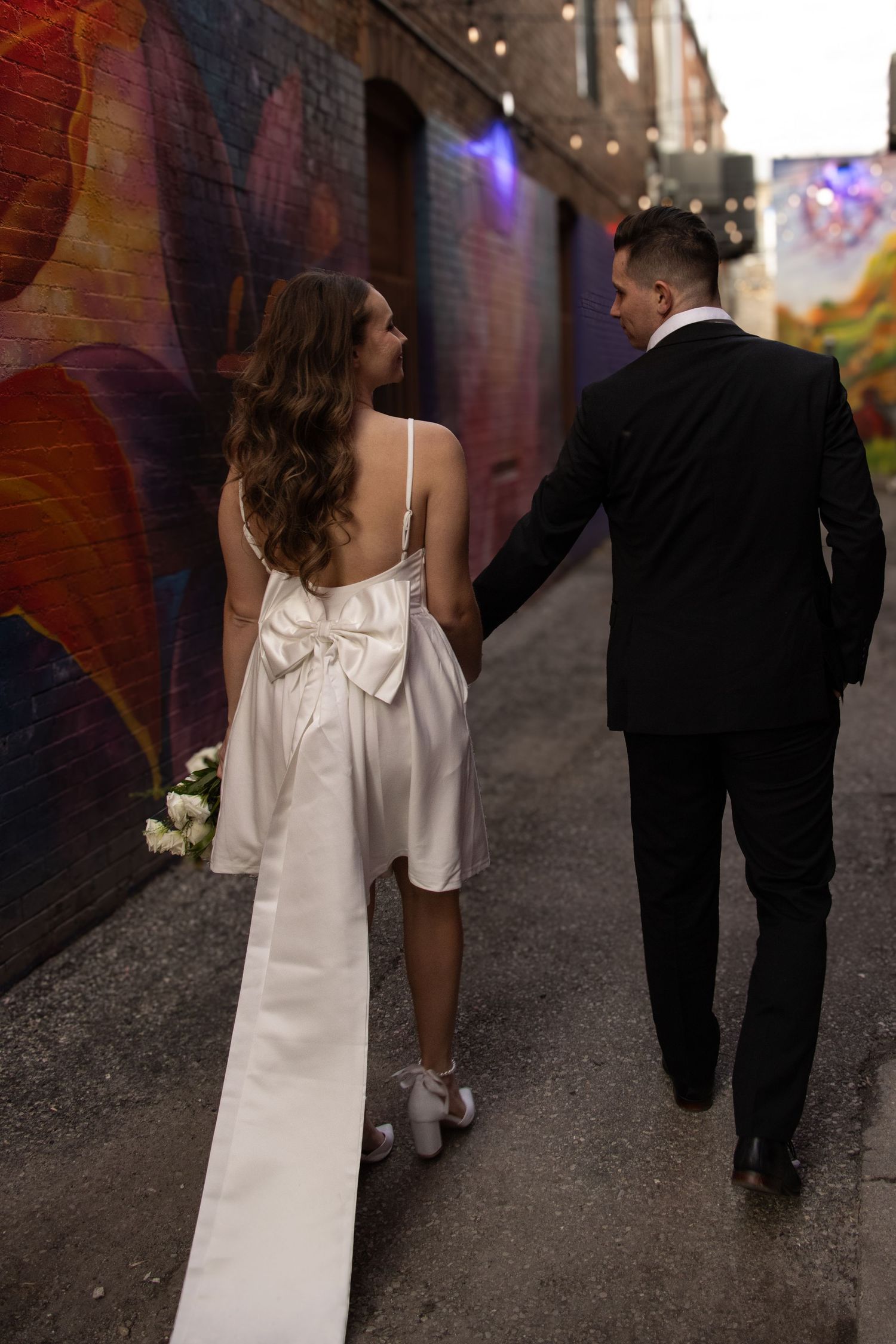 A couple dressed in formal attire walk hand-in-hand down a colorful, mural-lined alleyway. The woman wears a white dress