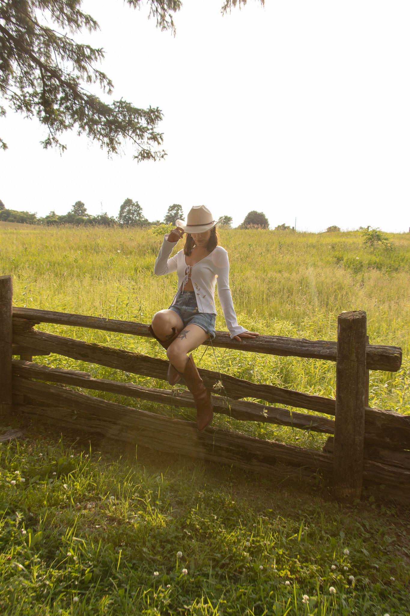 A person wearing a white shirt, denim shorts, brown boots, and a beige hat sits on a wooden fence amid a grassy field, h